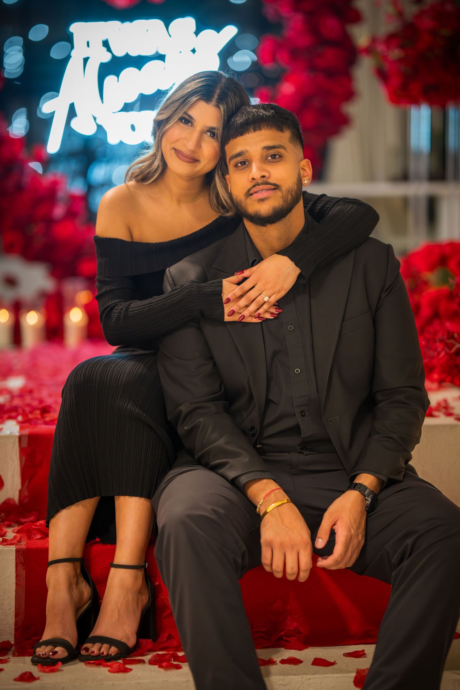 Couple in black formal wear poses intimately against red floral backdrop with neon sign.