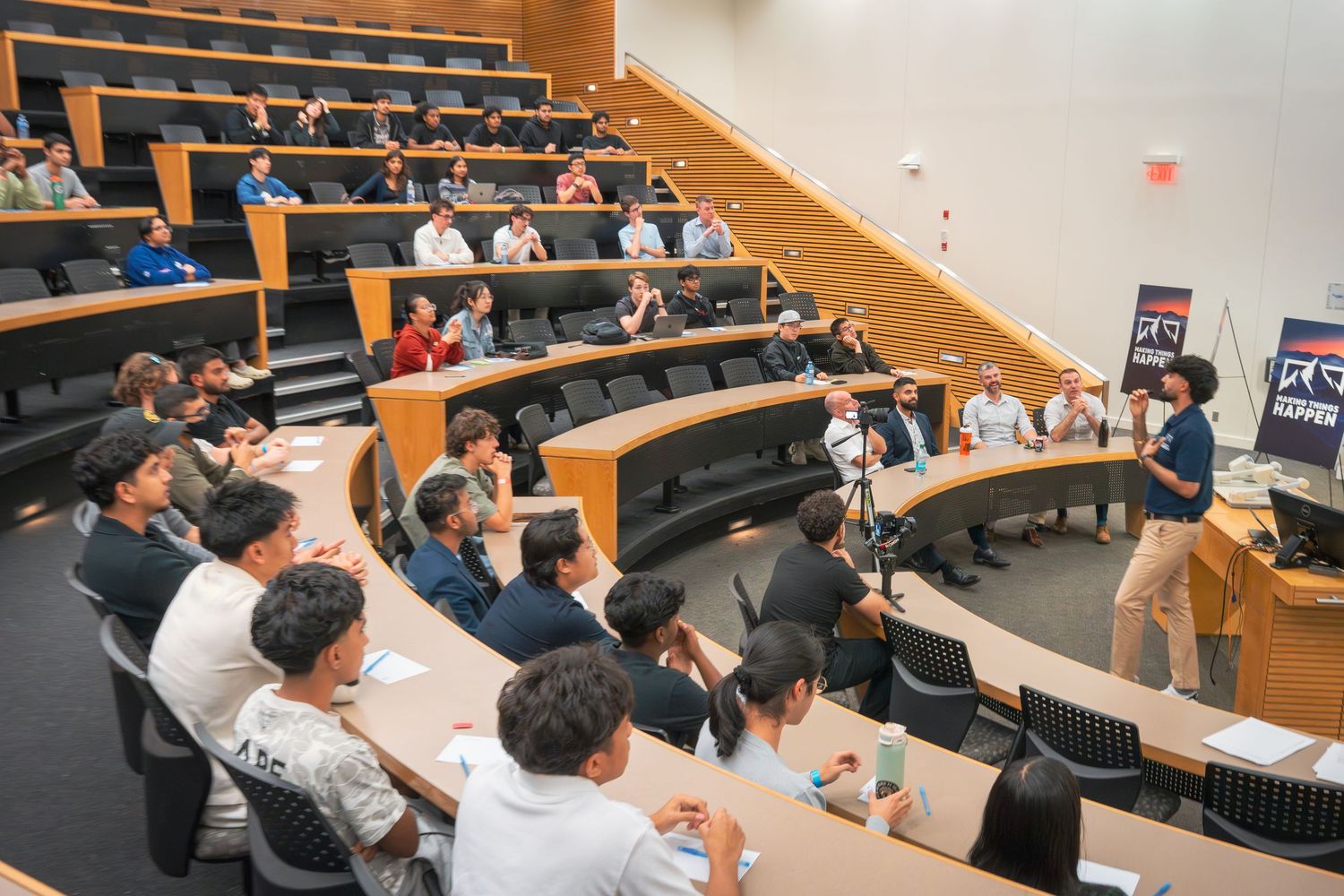 Business professionals attend a presentation in a modern conference room with tiered seating and wood paneling.