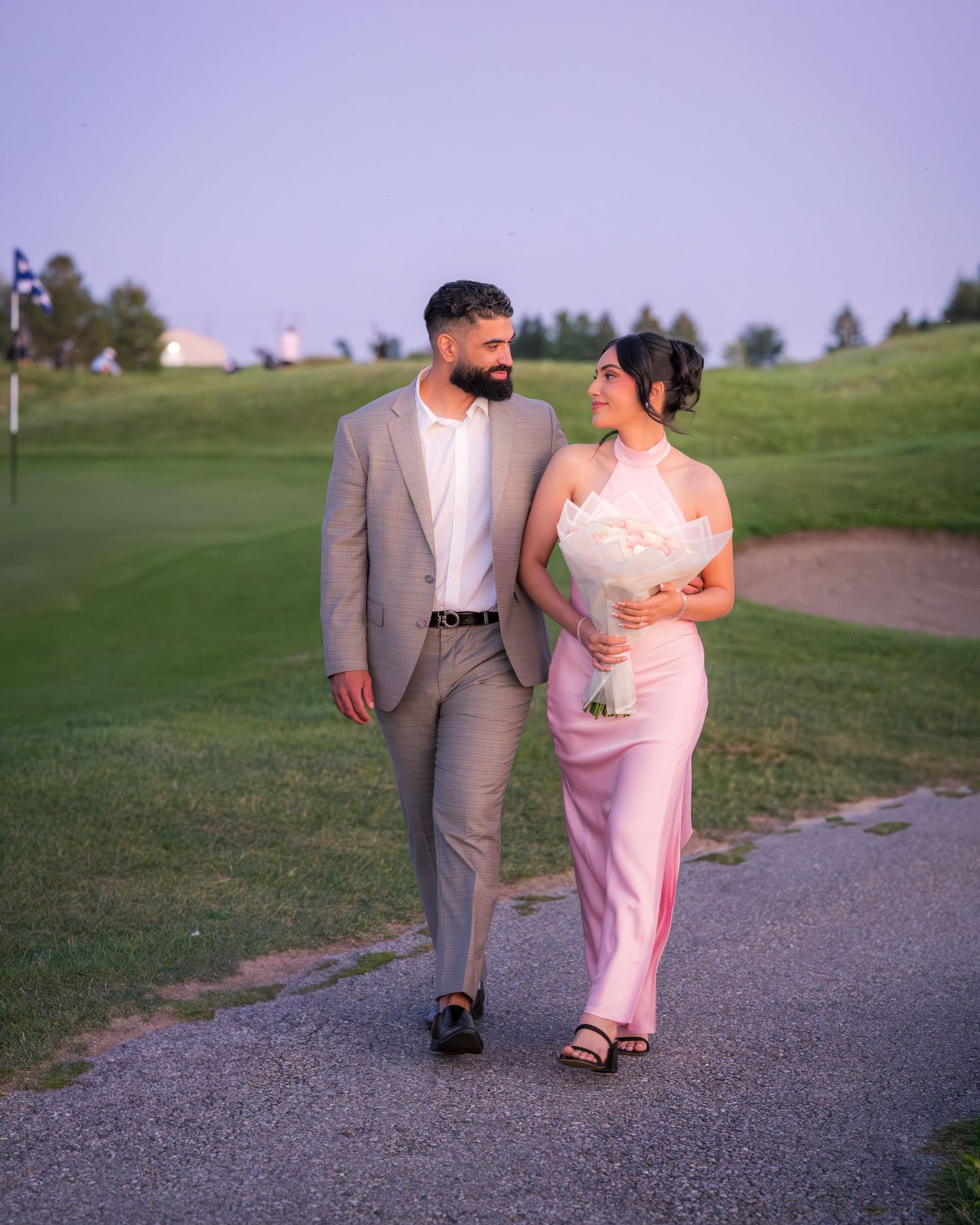 A couple strolls along a gravel path at sunset, wearing elegant pink and grey outfits.