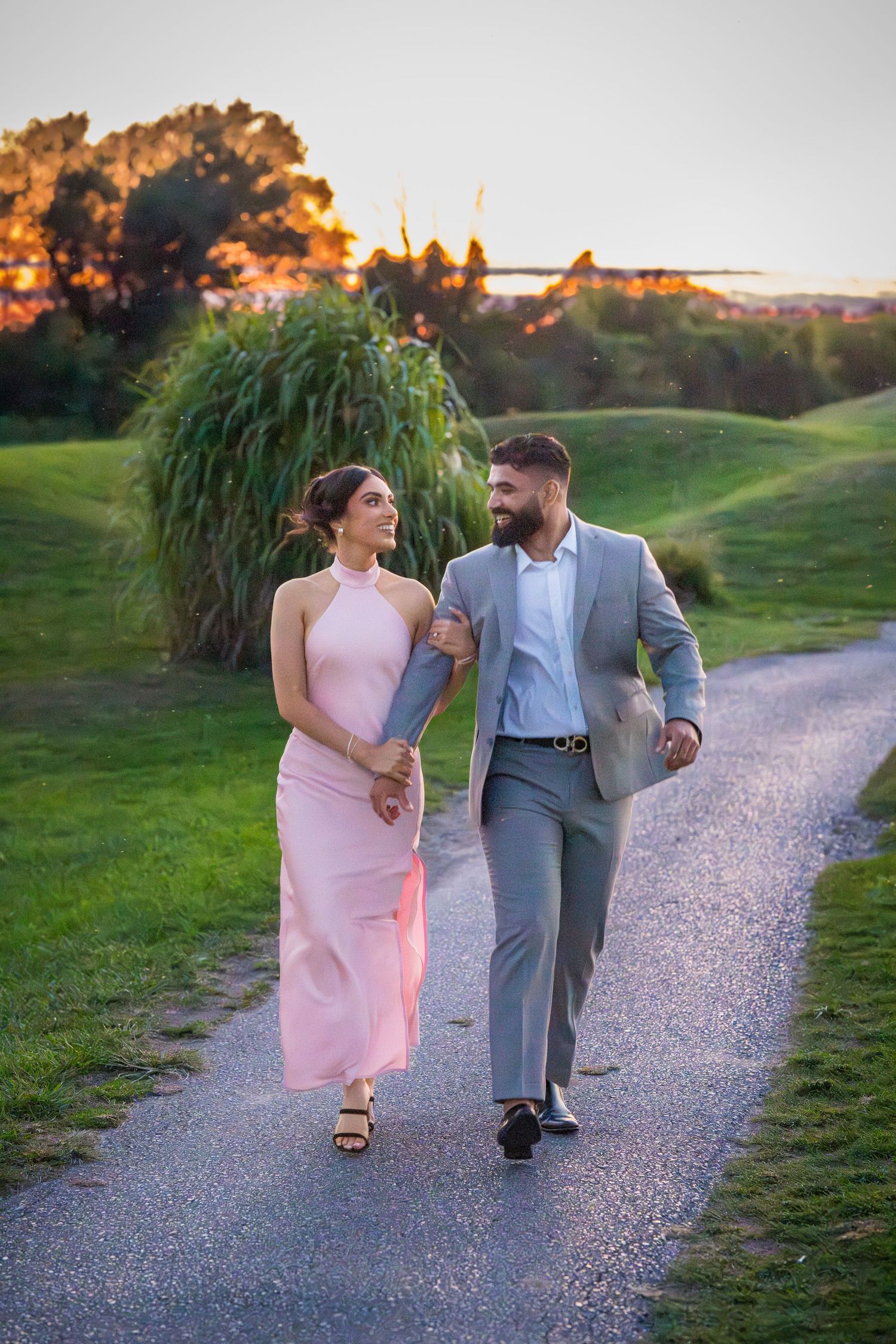 Two people in coordinated formal wear stroll together on an outdoor path at dusk.