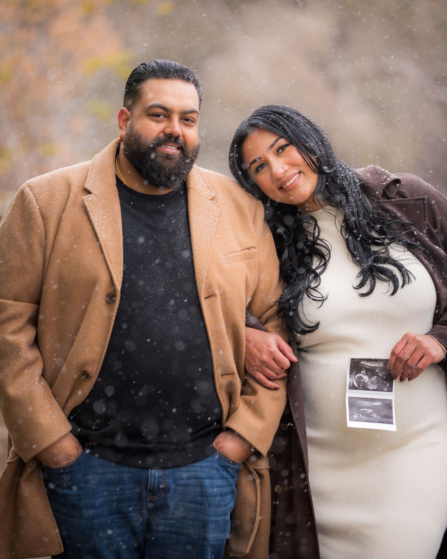 A couple poses together outdoors on a snowy day while holding a sonogram photo.