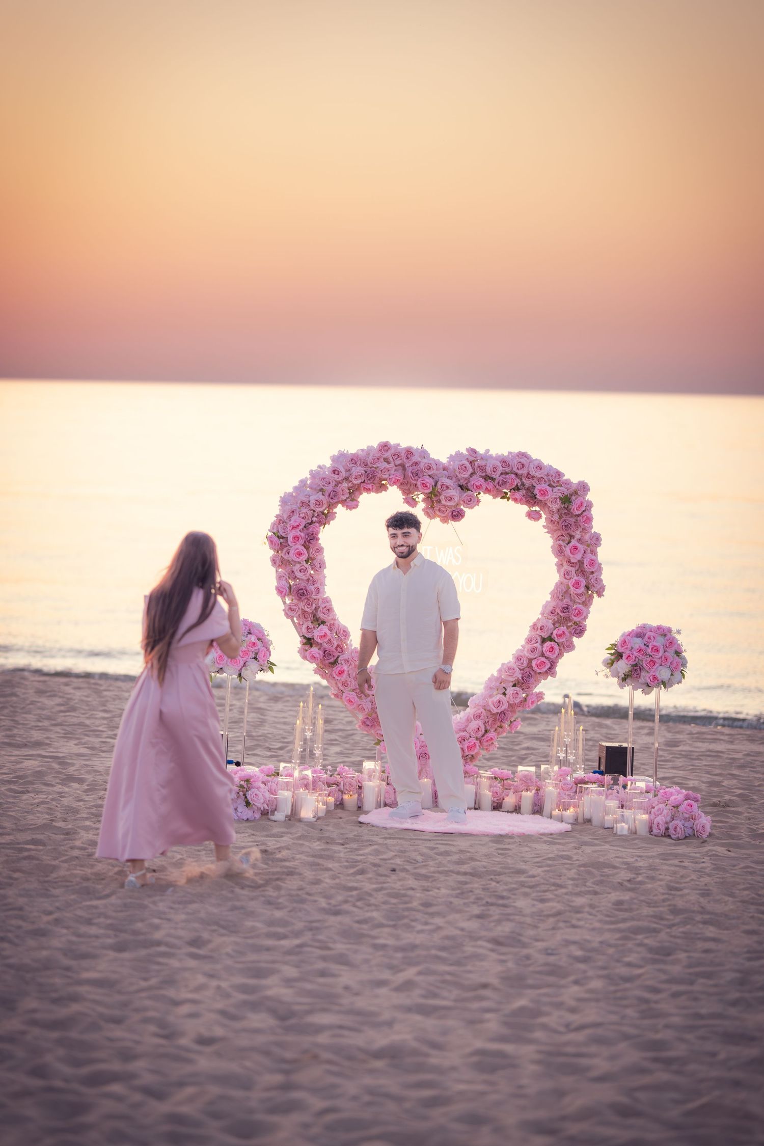 A beach proposal scene with a pink floral heart arch and lanterns during golden hour.