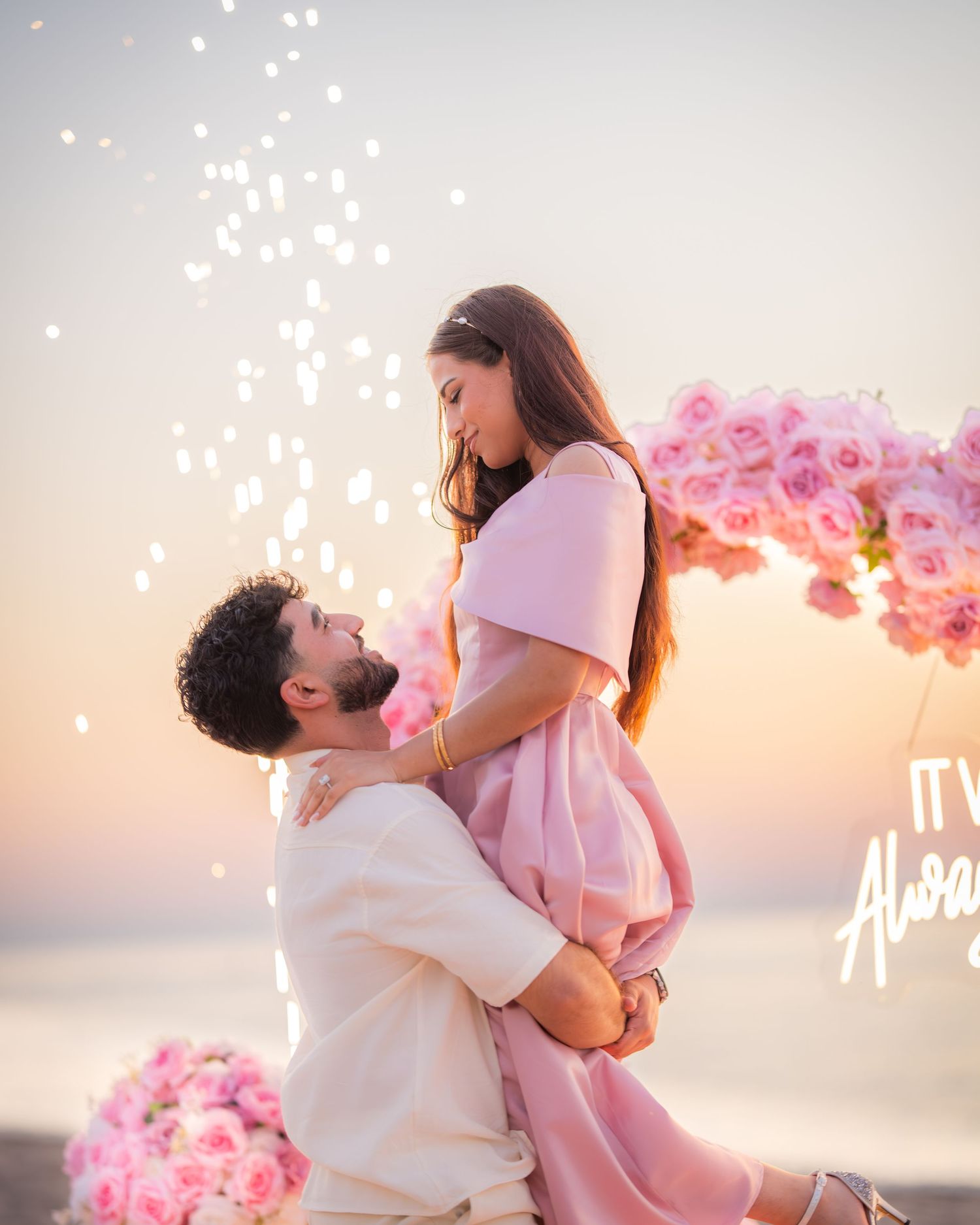 Magical beach proposal moment with sparklers, pink floral arch, and sunset backdrop over the ocean.
