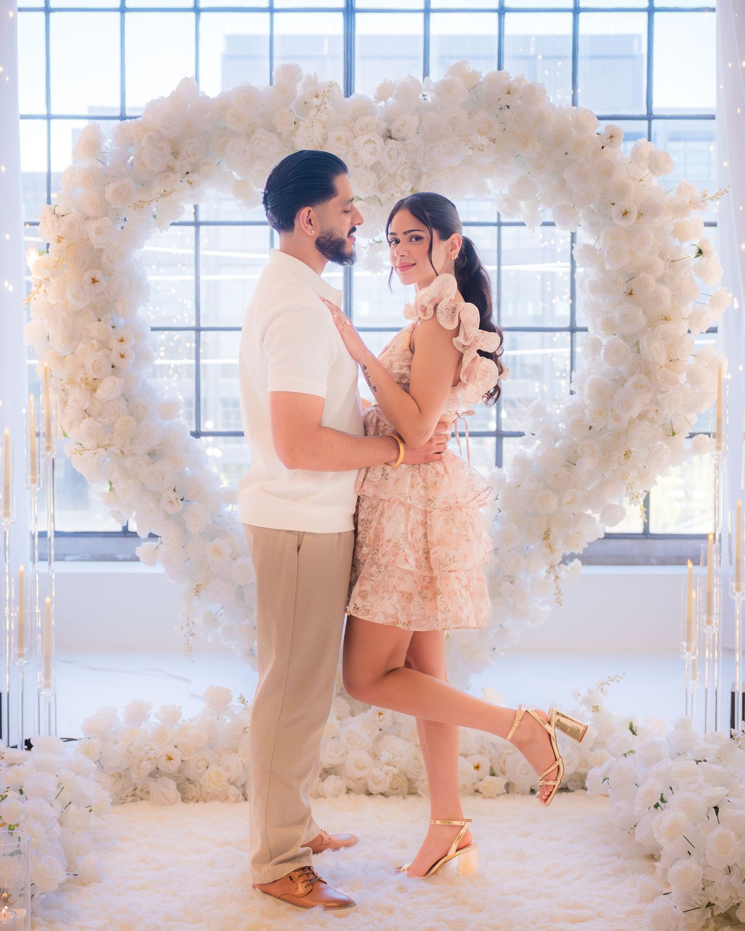 Couple posing romantically in front of a large white floral heart arch, woman in floral dress, man in white shirt.