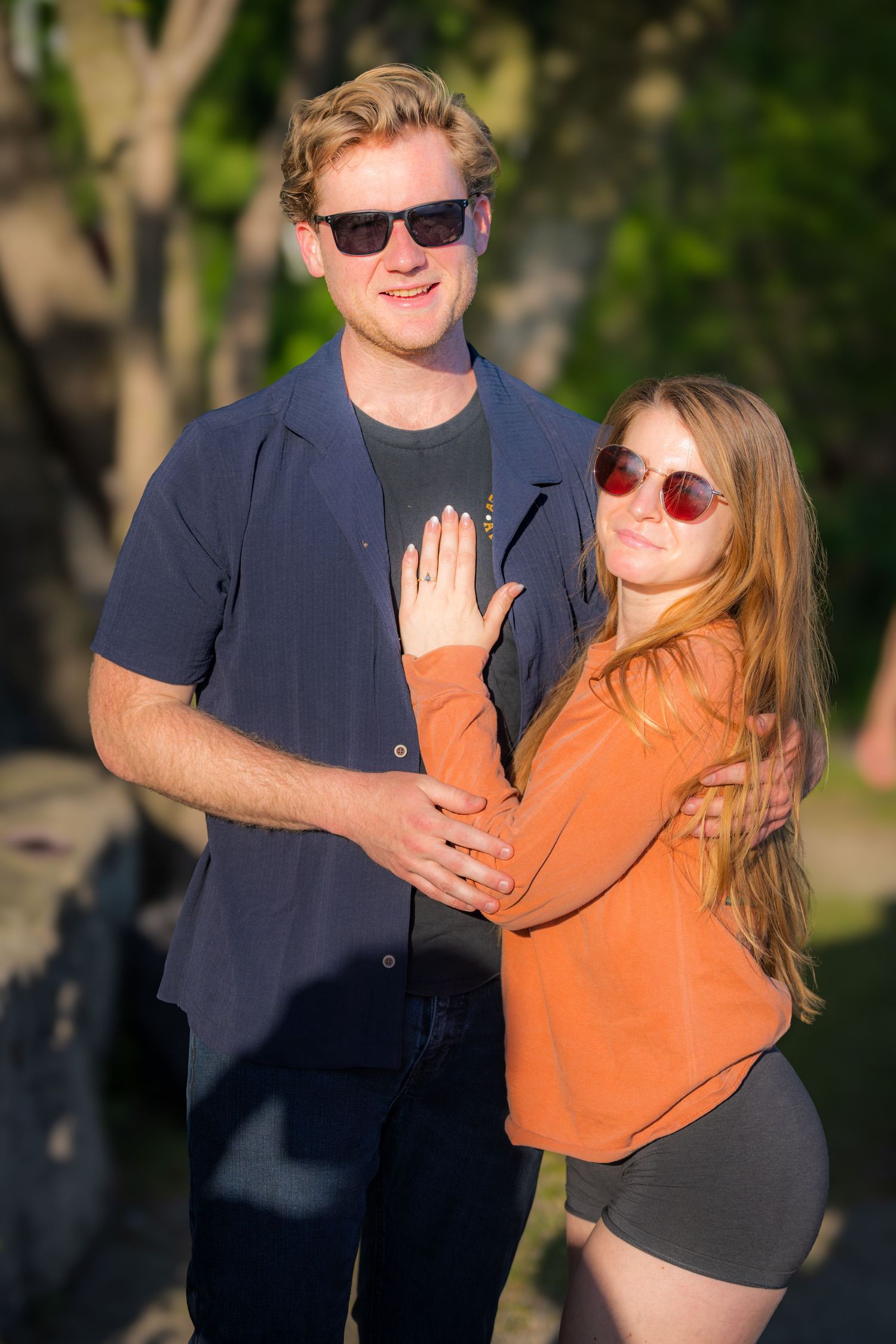 Young engaged couple posing outdoors in sunglasses, woman showing off her engagement ring.