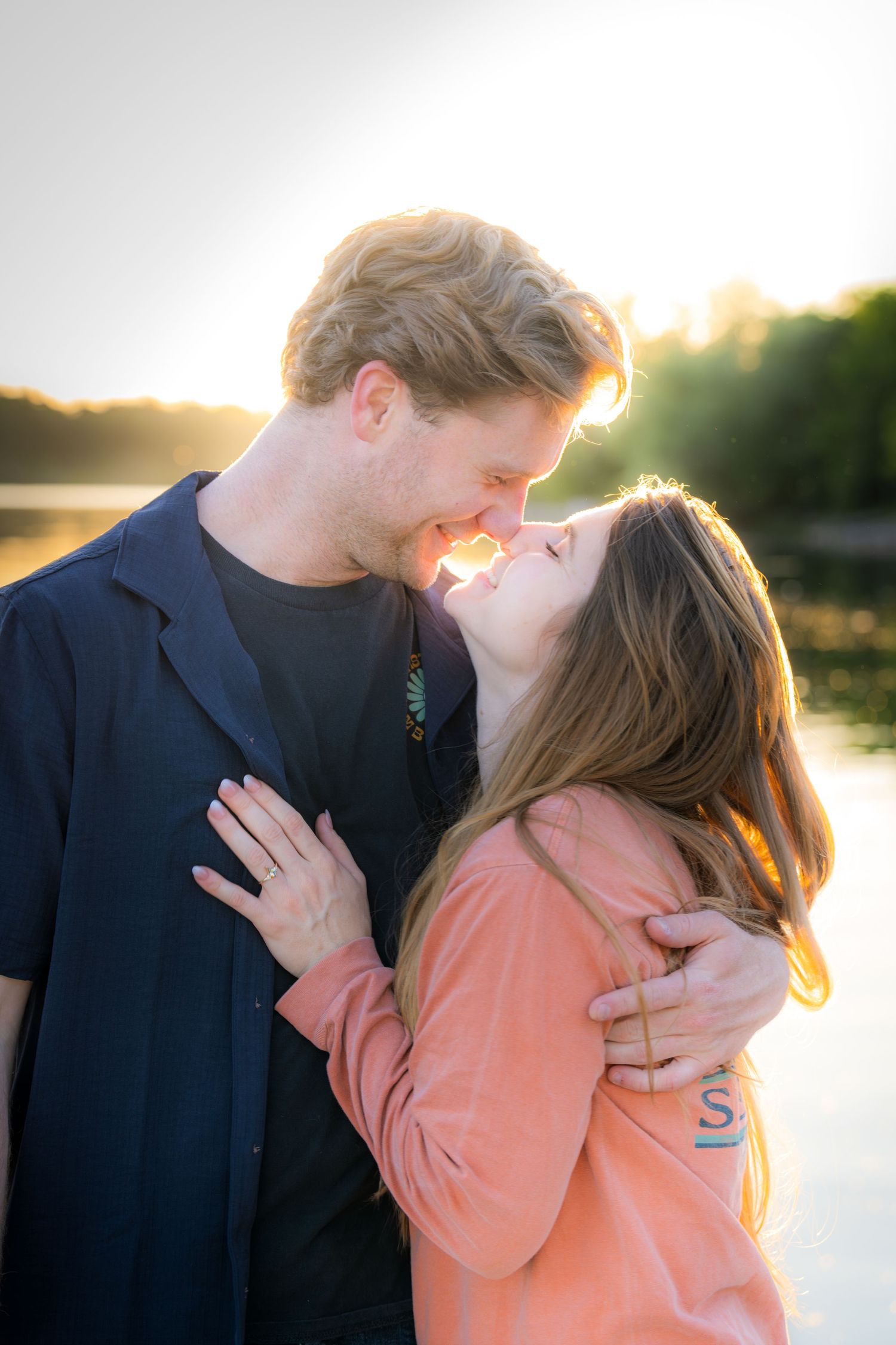 Couple sharing a tender kiss while embracing near a lake during a beautiful golden sunset.