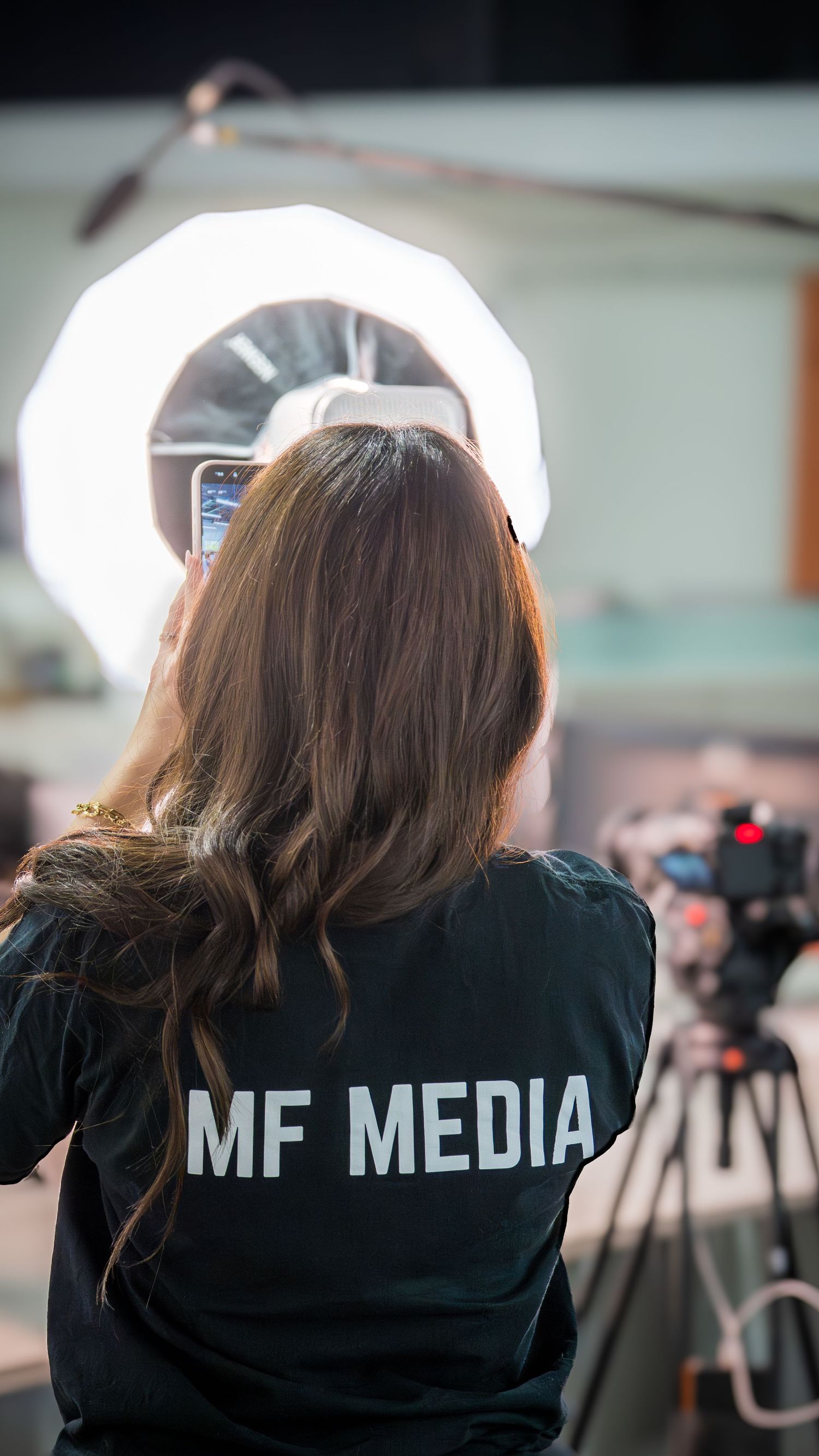 Woman in MF Media shirt holds phone near ring light and camera in professional media production studio.