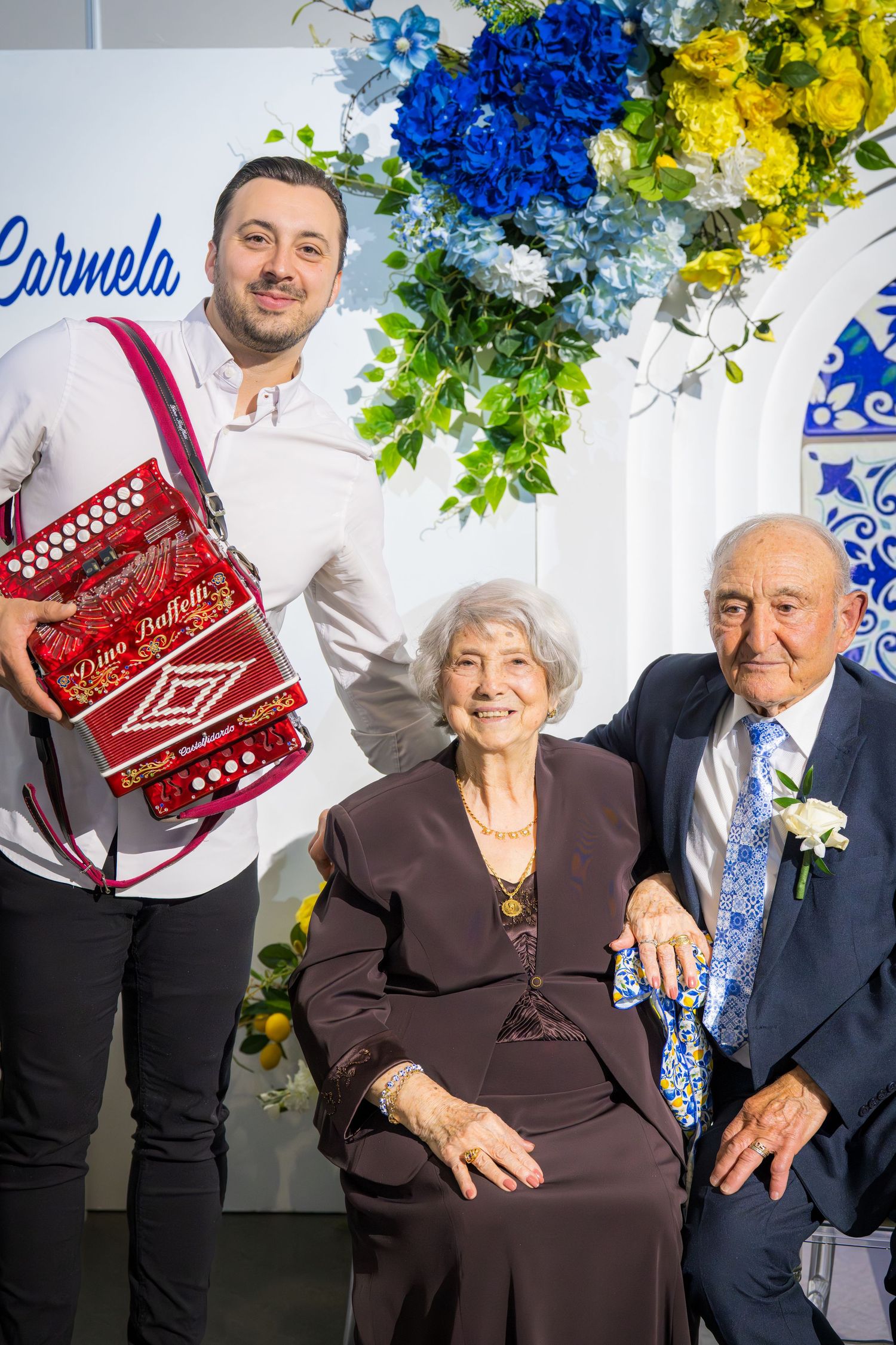 Accordion musician posing with elderly couple at Carmela-themed celebration with blue and yellow floral backdrop.