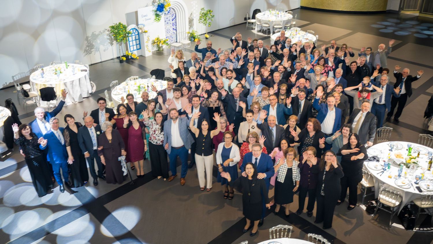 Large group of guests celebrate anniversary party in elegant venue with blue and white decor.