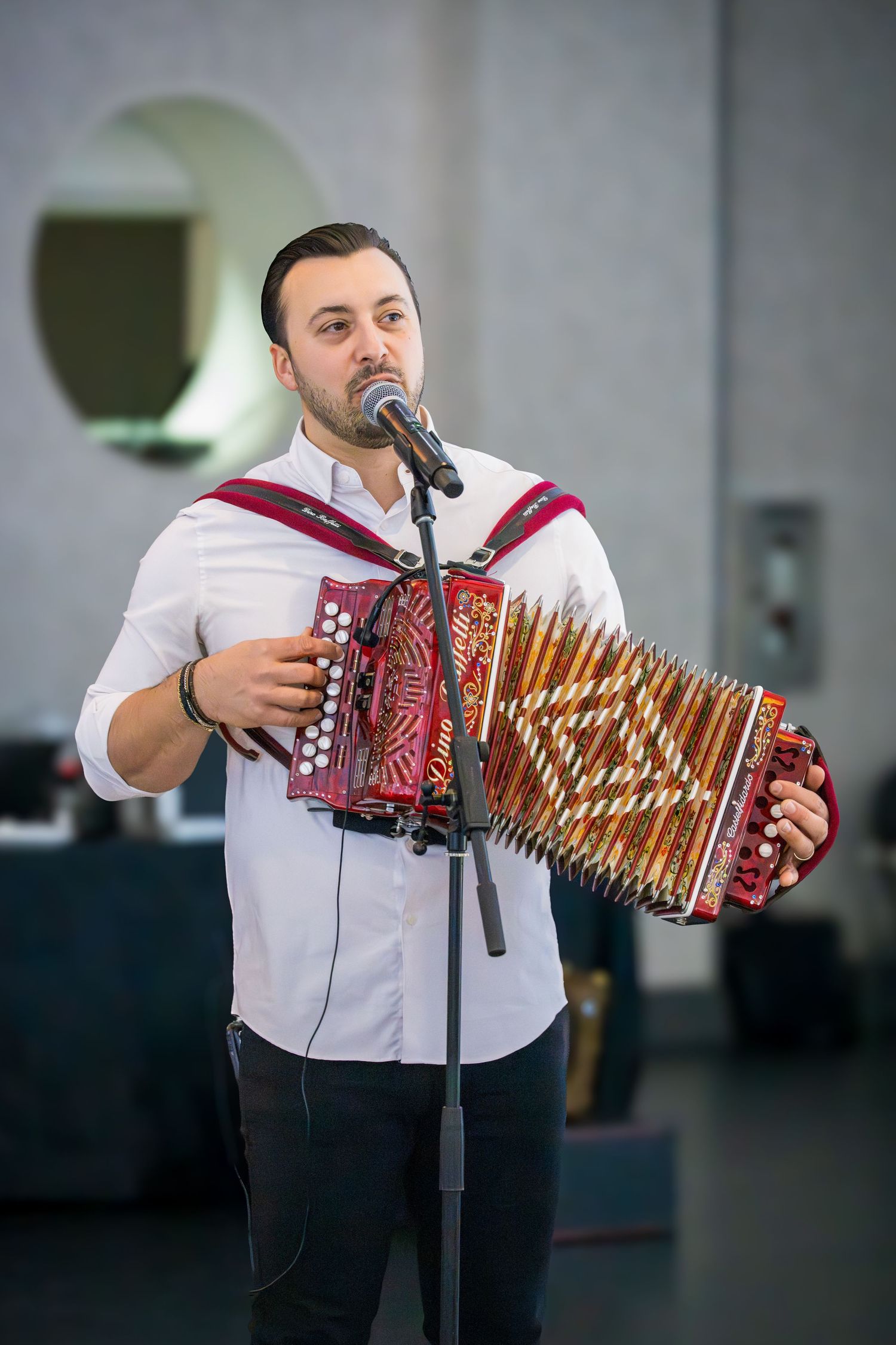 Man in white shirt playing red accordion and singing into microphone at live performance.