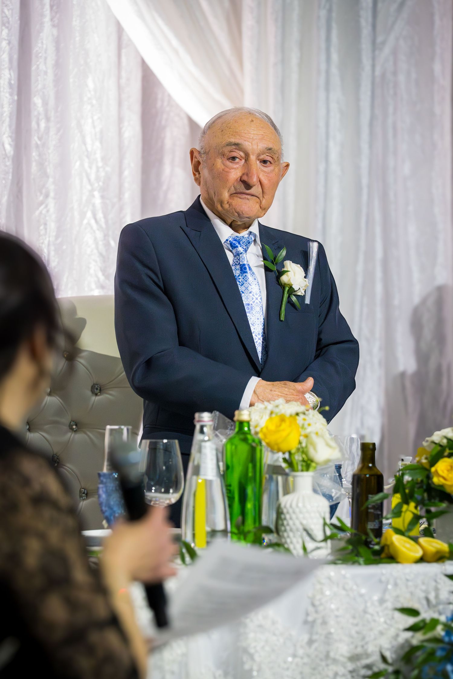 Elderly man in navy suit with floral boutonniere standing at a wedding reception head table.