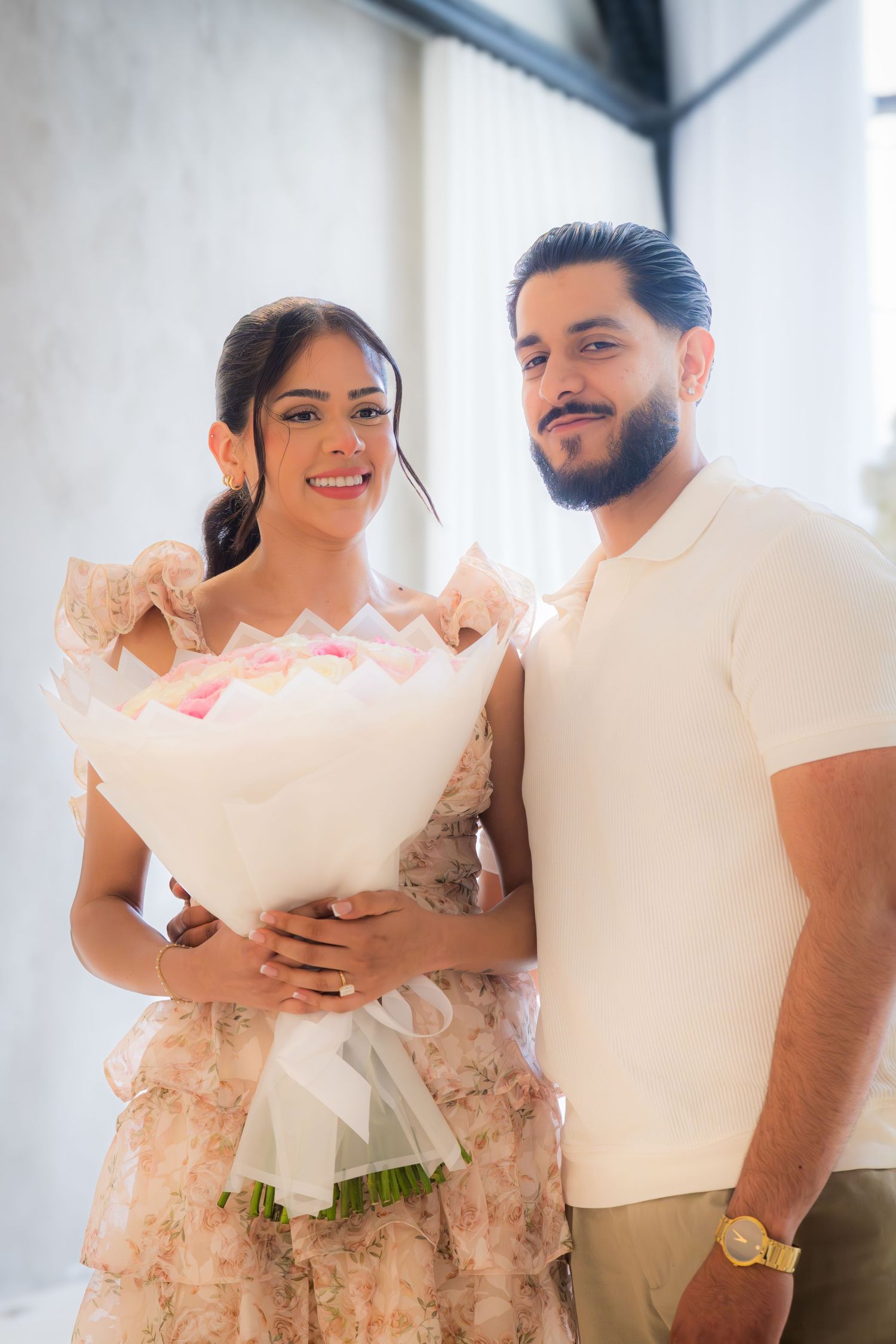 Happy couple posing together holding large bouquet of pink and white roses indoors.