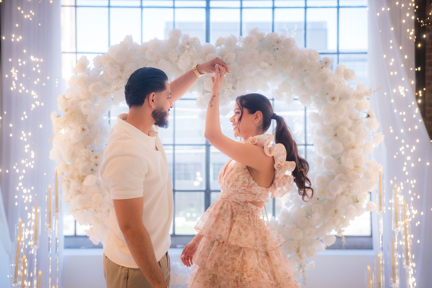 Man spinning woman in a floral dress under a white heart-shaped flower arch with lights.