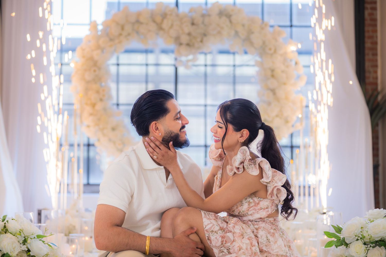 Couple sitting on the floor smiling at each other beneath a glowing white floral heart arch with fairy lights.