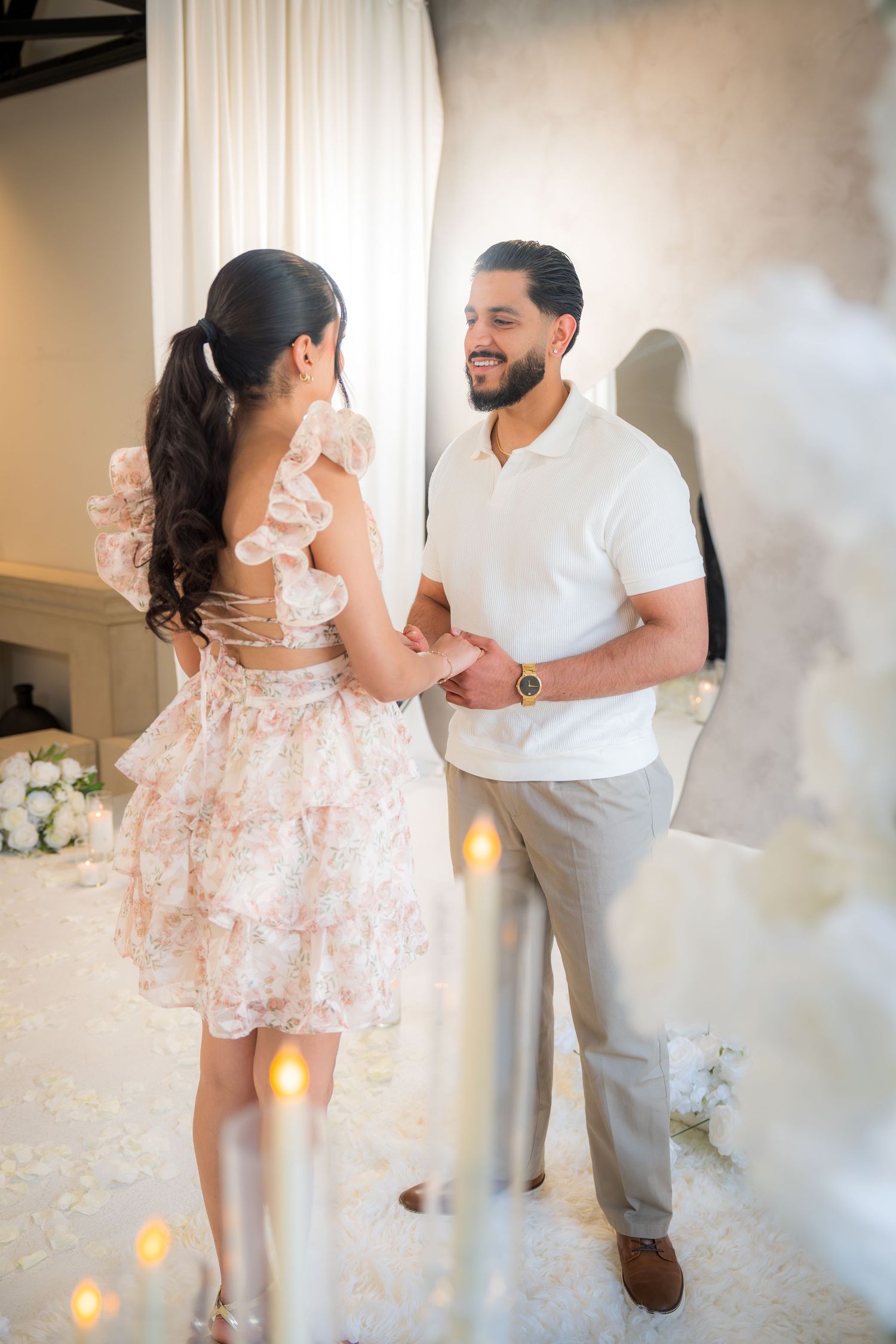 Smiling couple holding hands surrounded by candles and white floral decorations indoors.