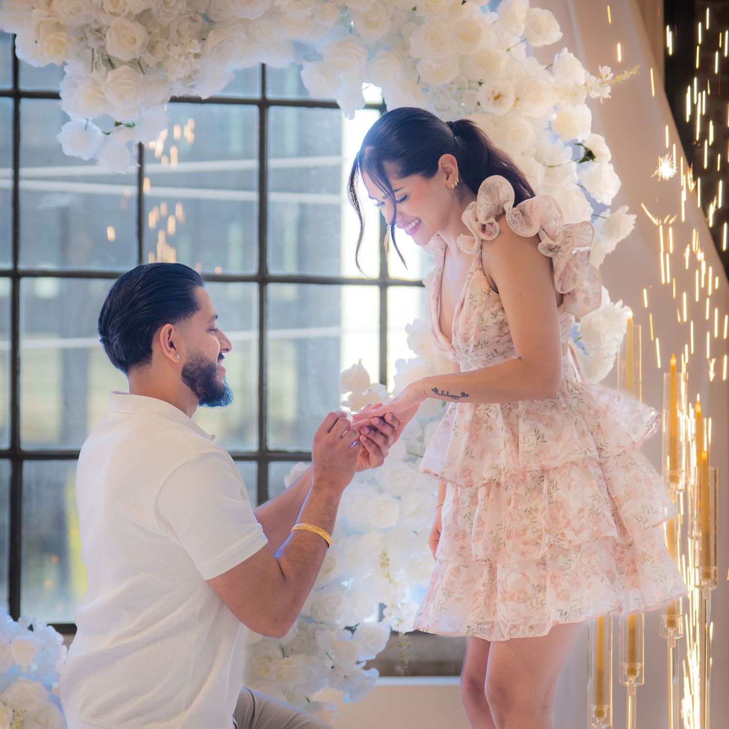 Man kneeling to propose with a ring to a surprised woman in a floral dress before a white flower arch.