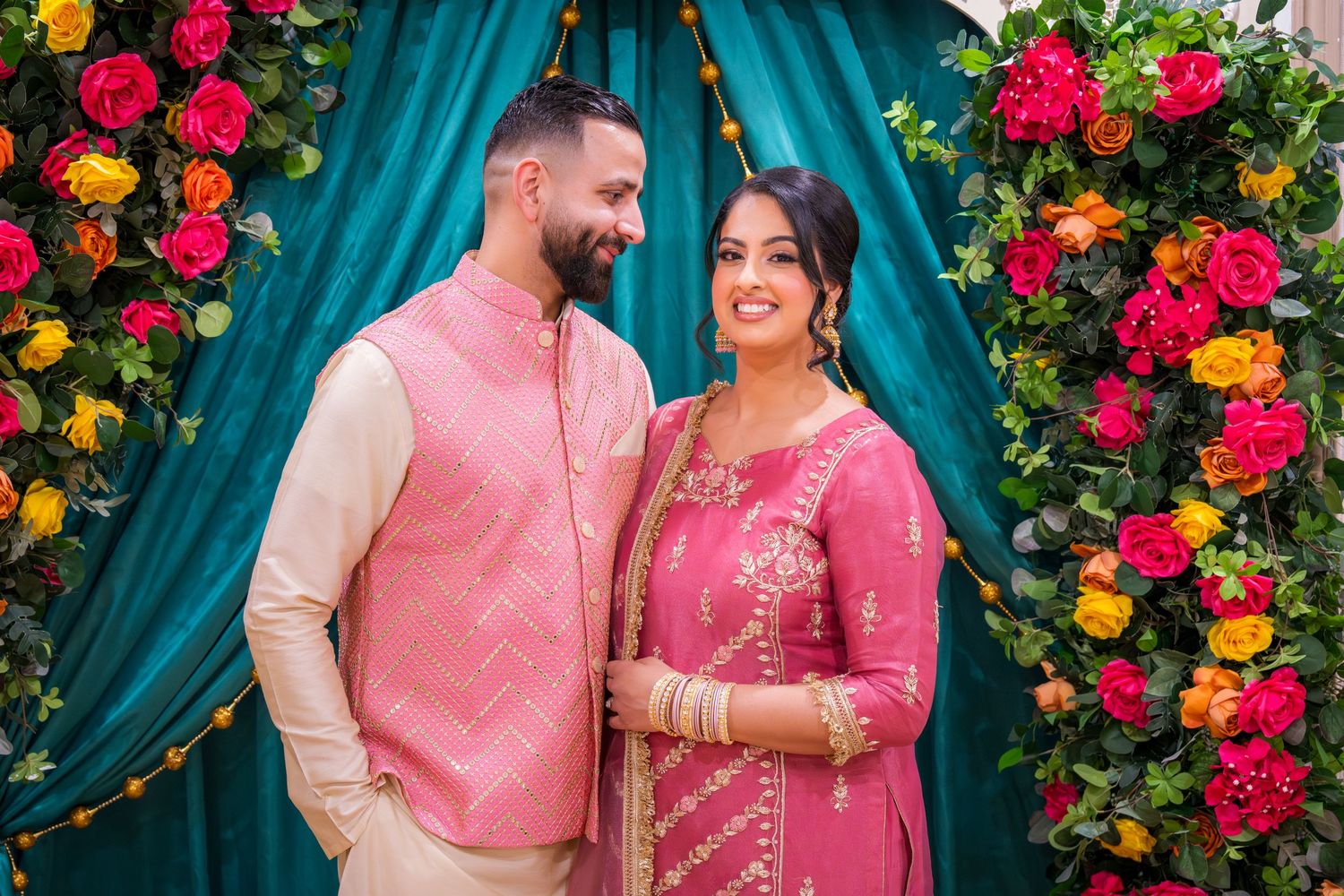 South Asian couple in pink traditional attire smiling together at colorful floral backdrop.