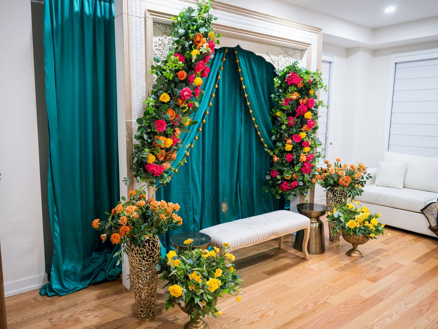 Elegant teal drape backdrop with vibrant floral arch and yellow roses in gold vases on hardwood floor.