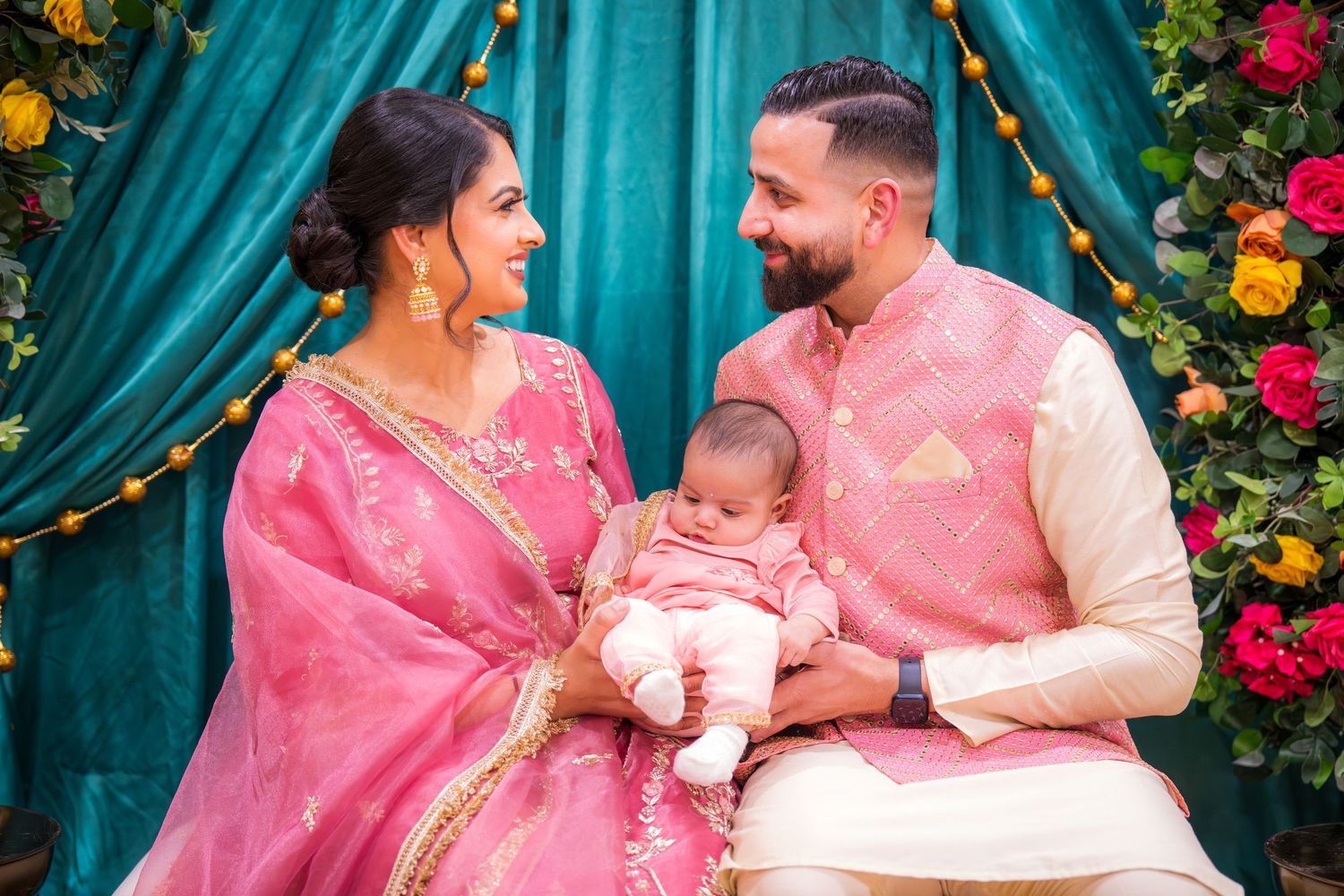 Indian couple in pink traditional attire sits together holding newborn at colorful floral teal backdrop.