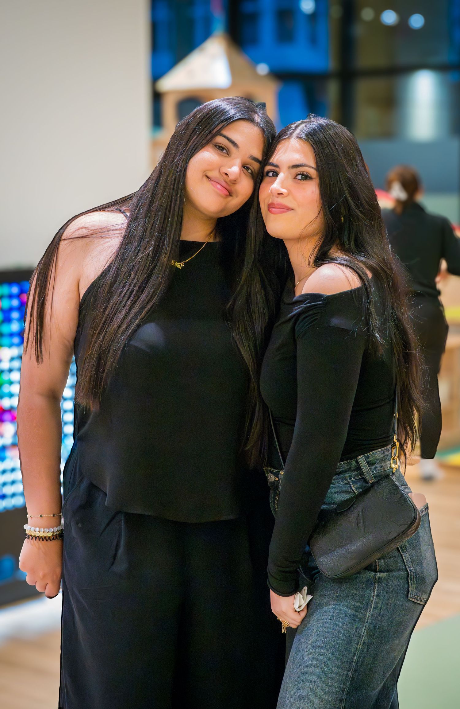 Two young women with long dark hair wearing black outfits pose together smiling at the camera.