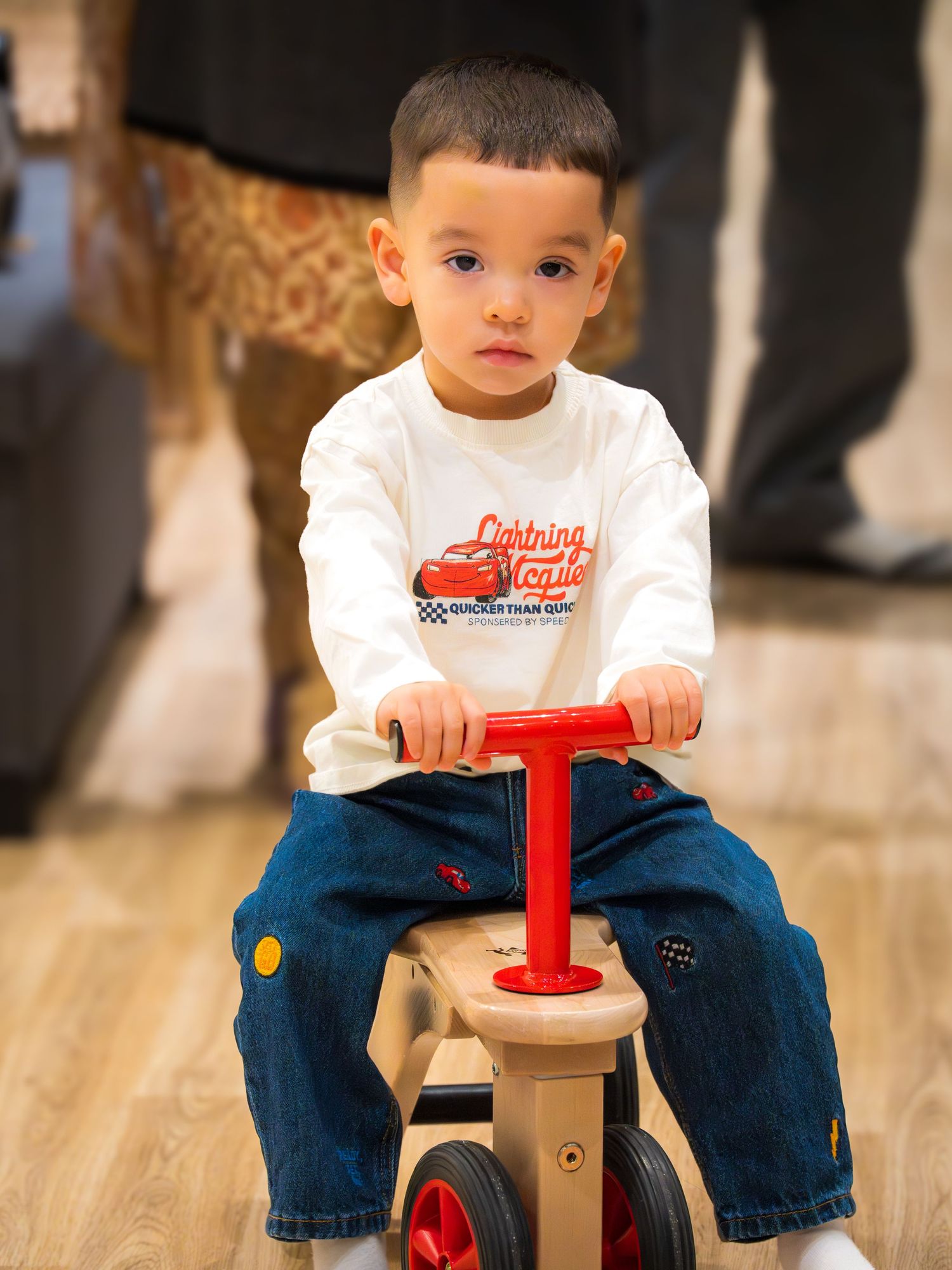 Young boy in Lightning McQueen shirt riding a wooden toy tricycle indoors.