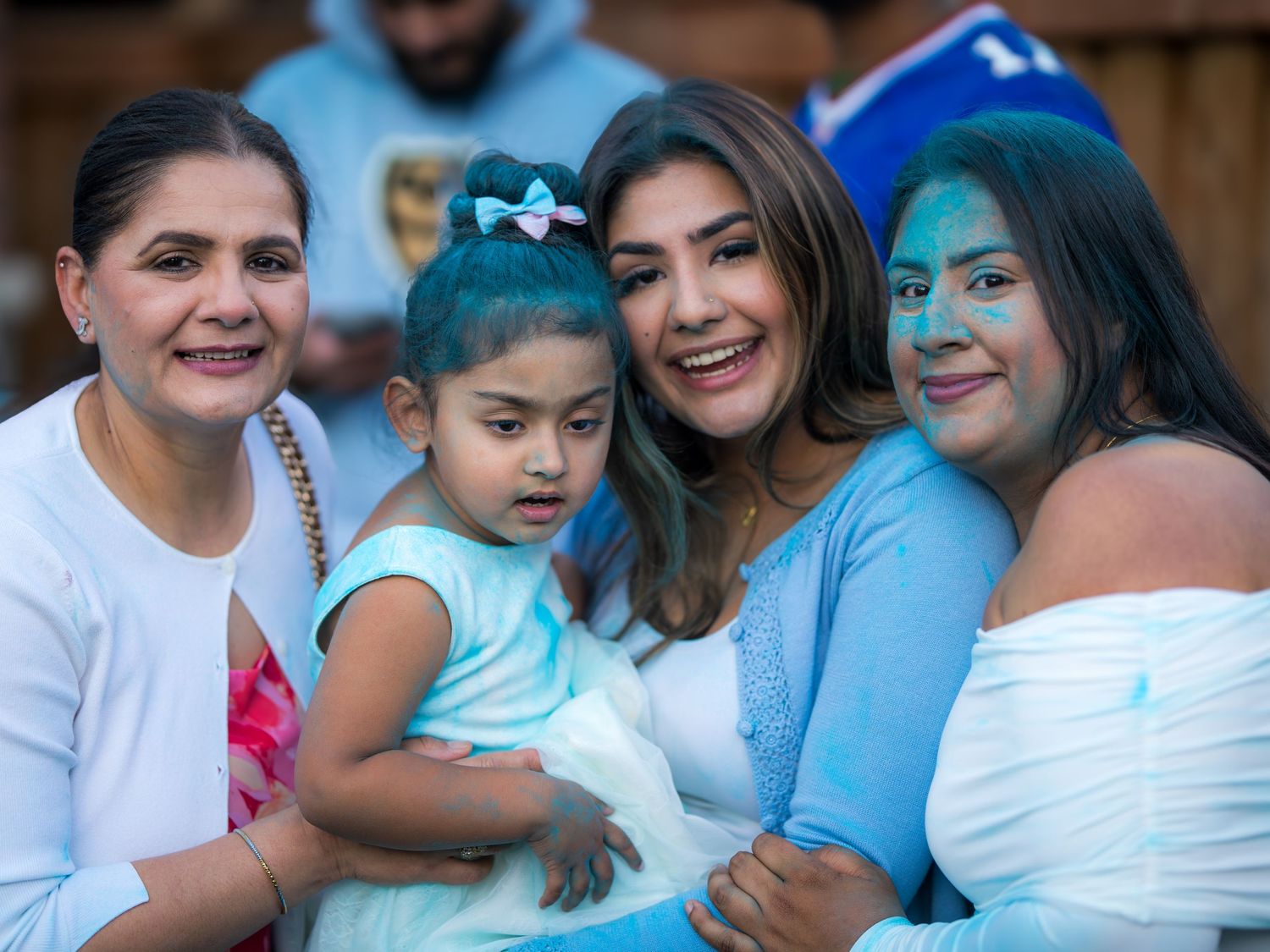 Four females including a toddler smile together with blue gender reveal powder at an outdoor party.