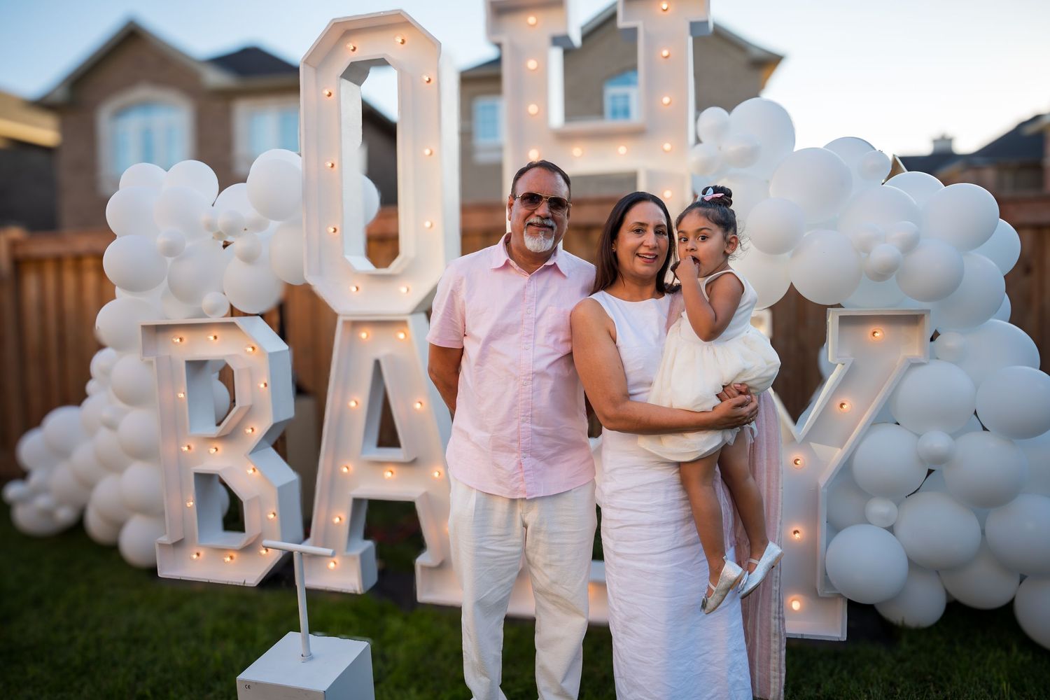 Man and woman holding young girl pose in front of lit OH BABY sign with white balloon decorations.