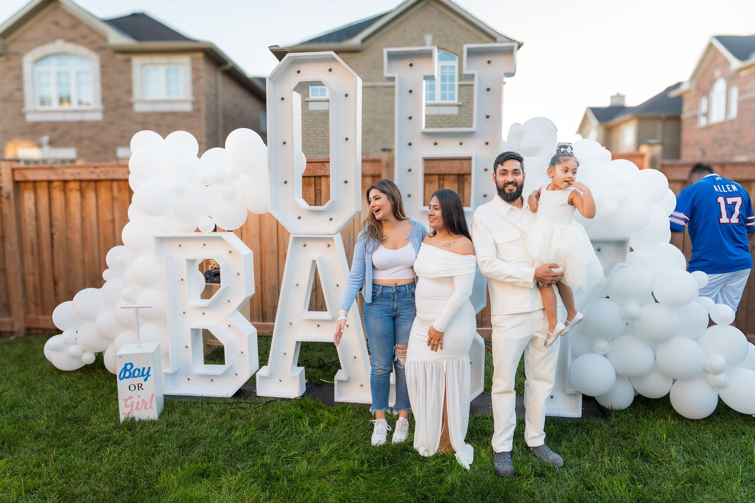 Family group poses in front of illuminated OH BABY marquee letters with white balloon arch at gender reveal.