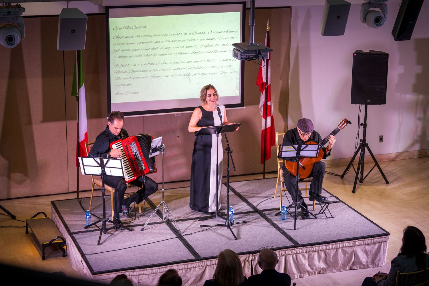 Female singer performs on stage with accordion and guitar duo beneath Italian and Canadian flags.