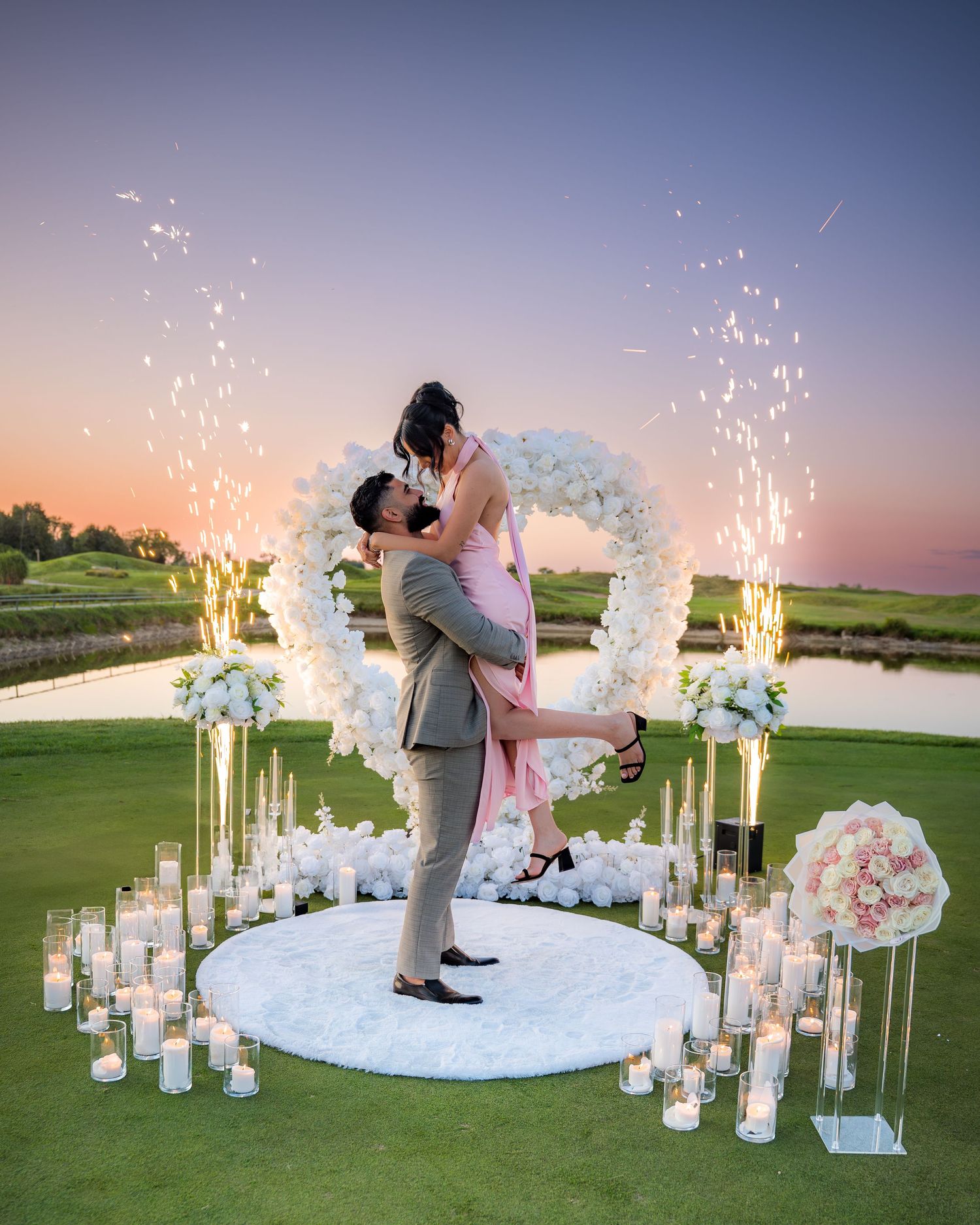 Couple sharing romantic moment under floral arch with sparklers at sunset.