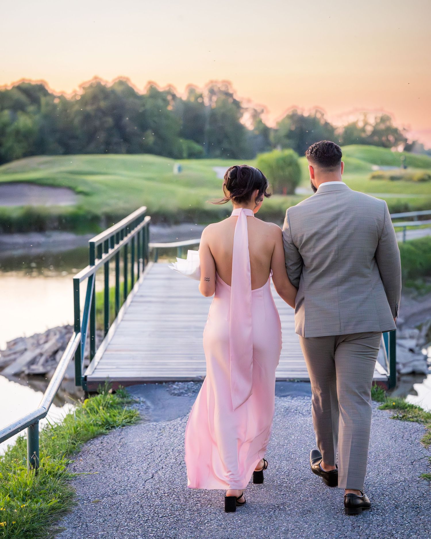 A couple in pink and gray formal wear walking on a path near a golf course at dusk.