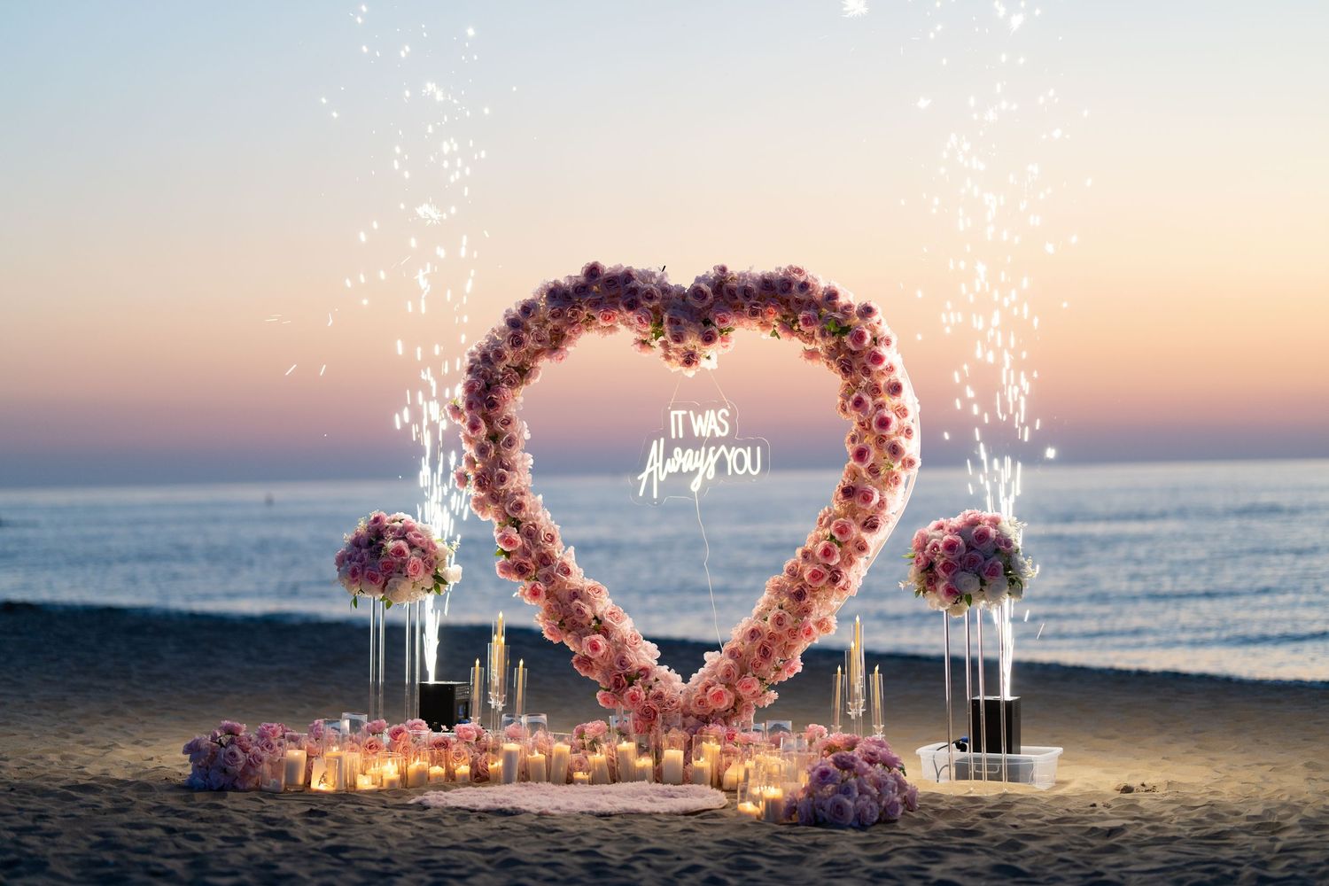 Pink floral heart arch and candle decorations on beach at golden hour.