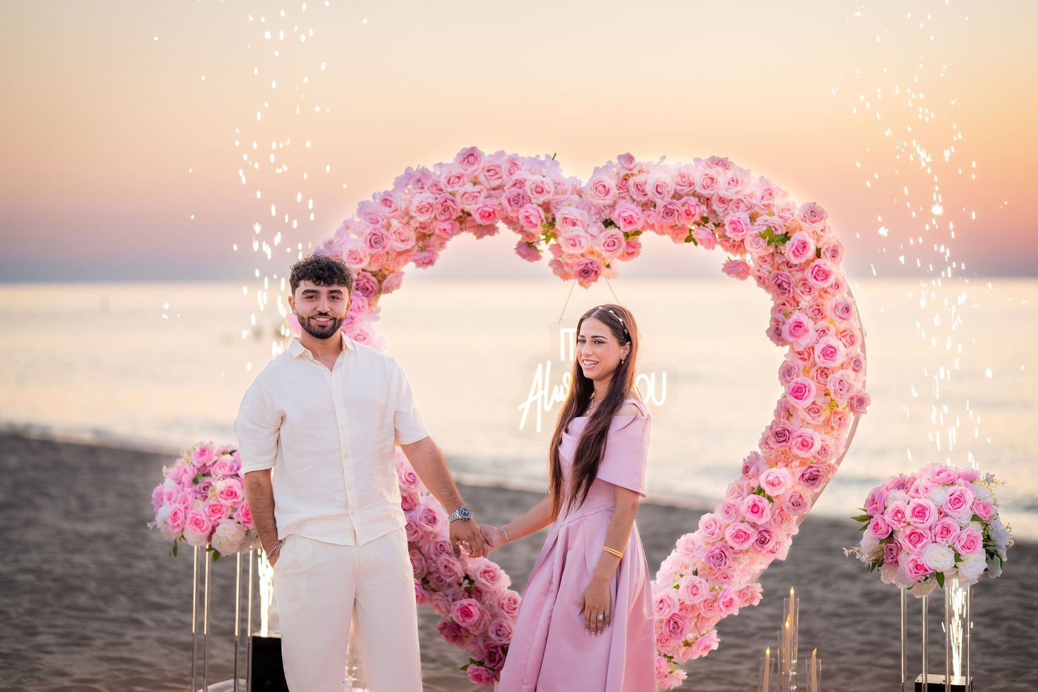 Purple and pink floral heart arch installation on sandy beach during golden hour.