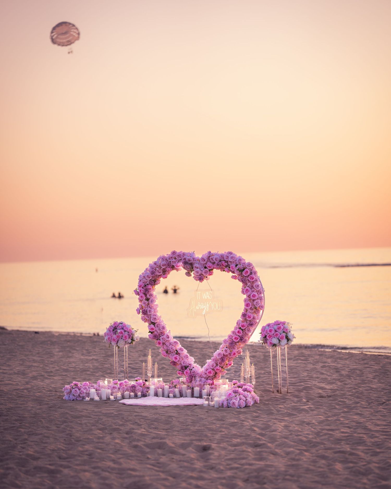 Heart-shaped floral arch on beach at golden hour with ocean backdrop.