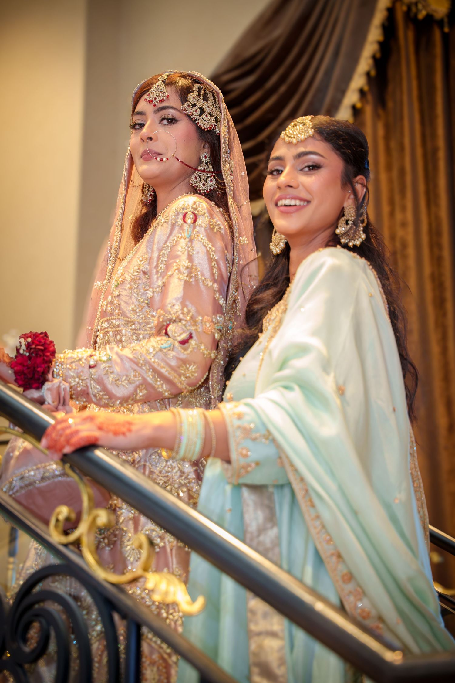 Two women in traditional South Asian attire pose elegantly on ornate staircase.