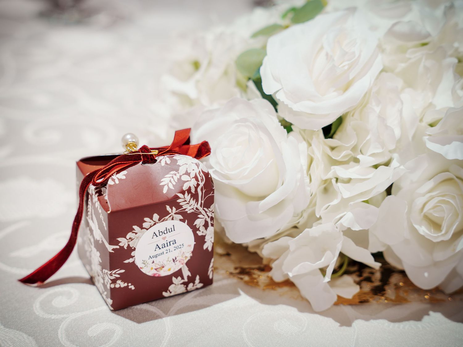 Elegant wedding favor box with white roses and red ribbon against light fabric background.