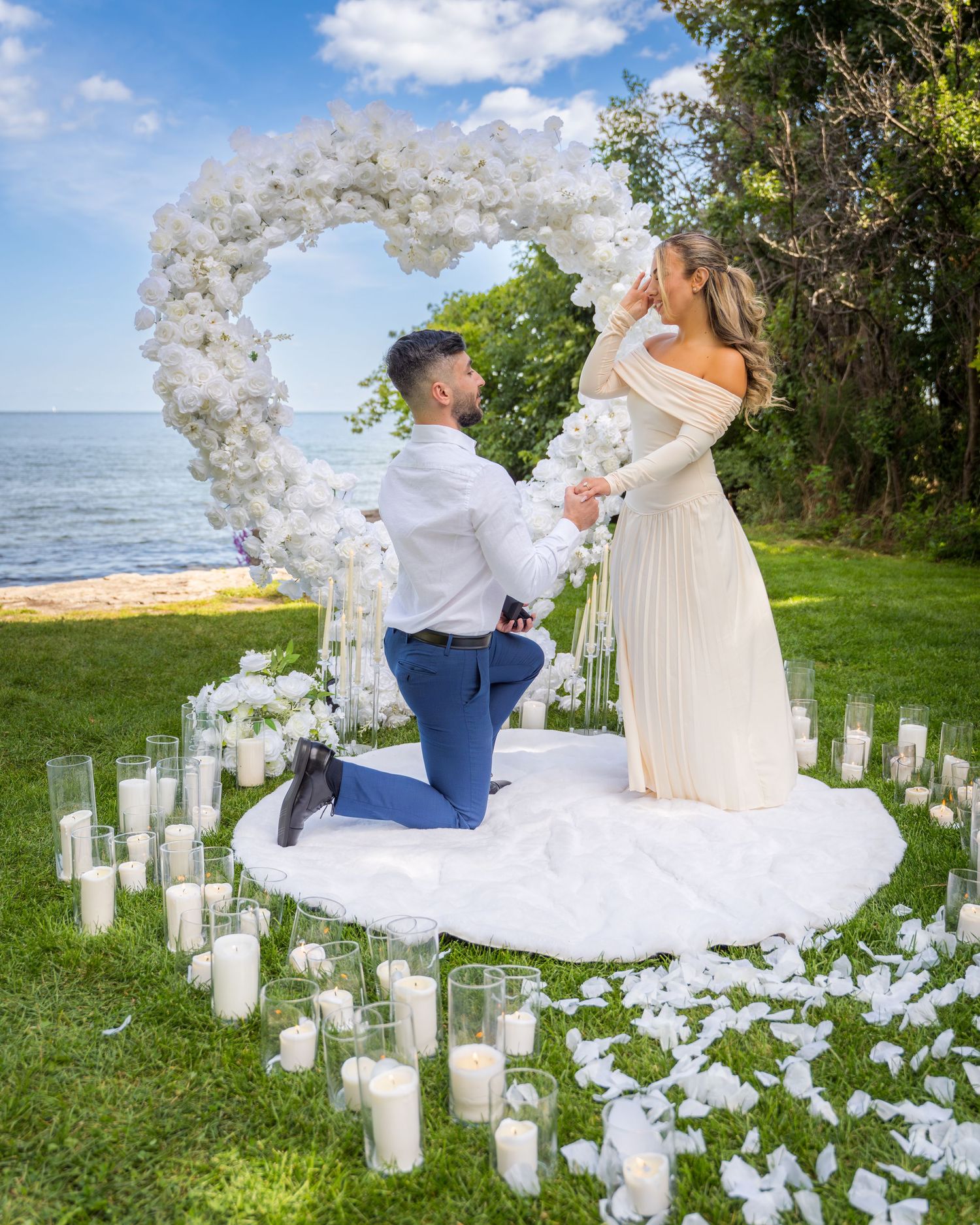 Proposal scene with white balloon arch and glowing candles on oceanfront lawn.