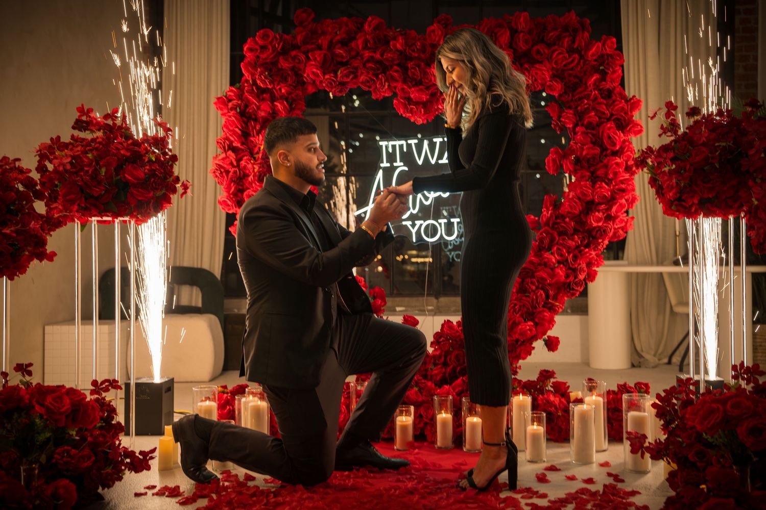 Romantic proposal setup with red flower wall, candles, and neon signage.
