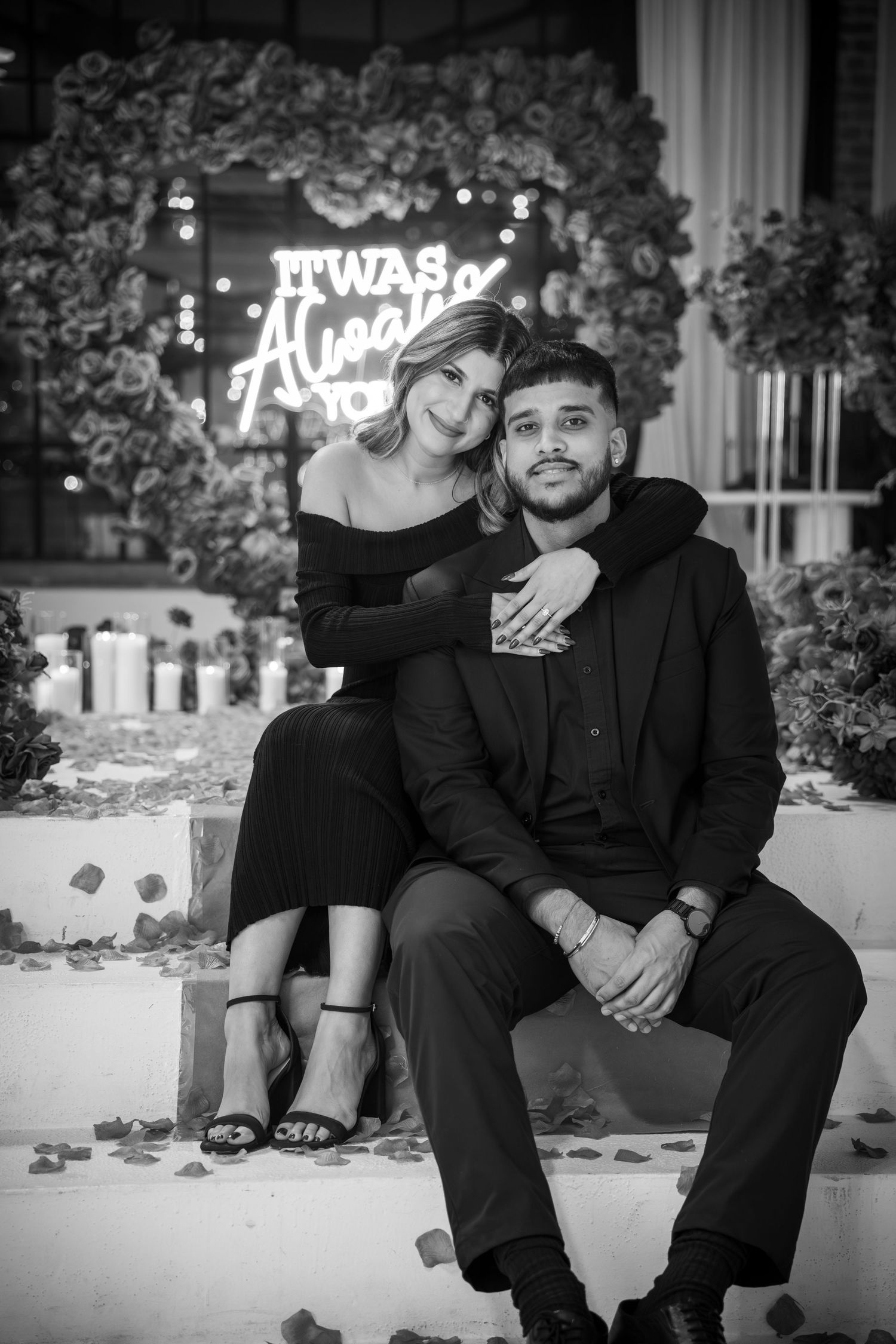 Couple sitting together on white ledge with neon sign and floral backdrop.