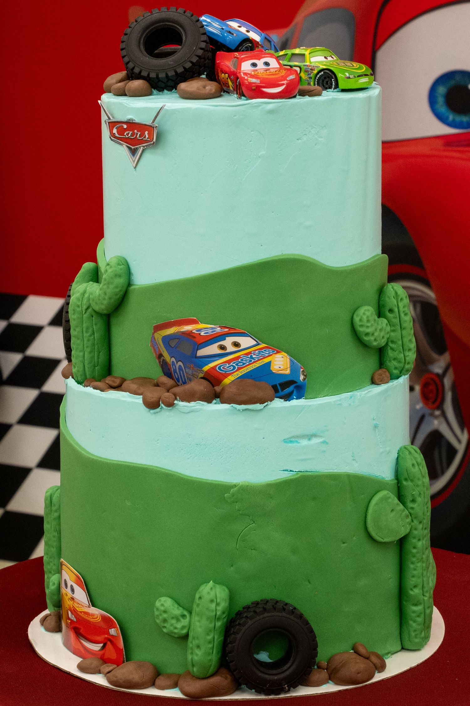 Three-tier Cars movie themed birthday cake with Lightning McQueen decorations, cacti, and toy cars on light blue and green fondant.
