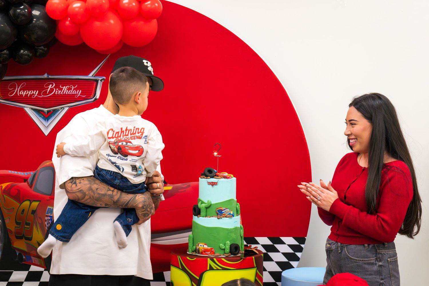 A woman claps as a tattooed man holds a child near a Cars-themed birthday cake with red balloons.