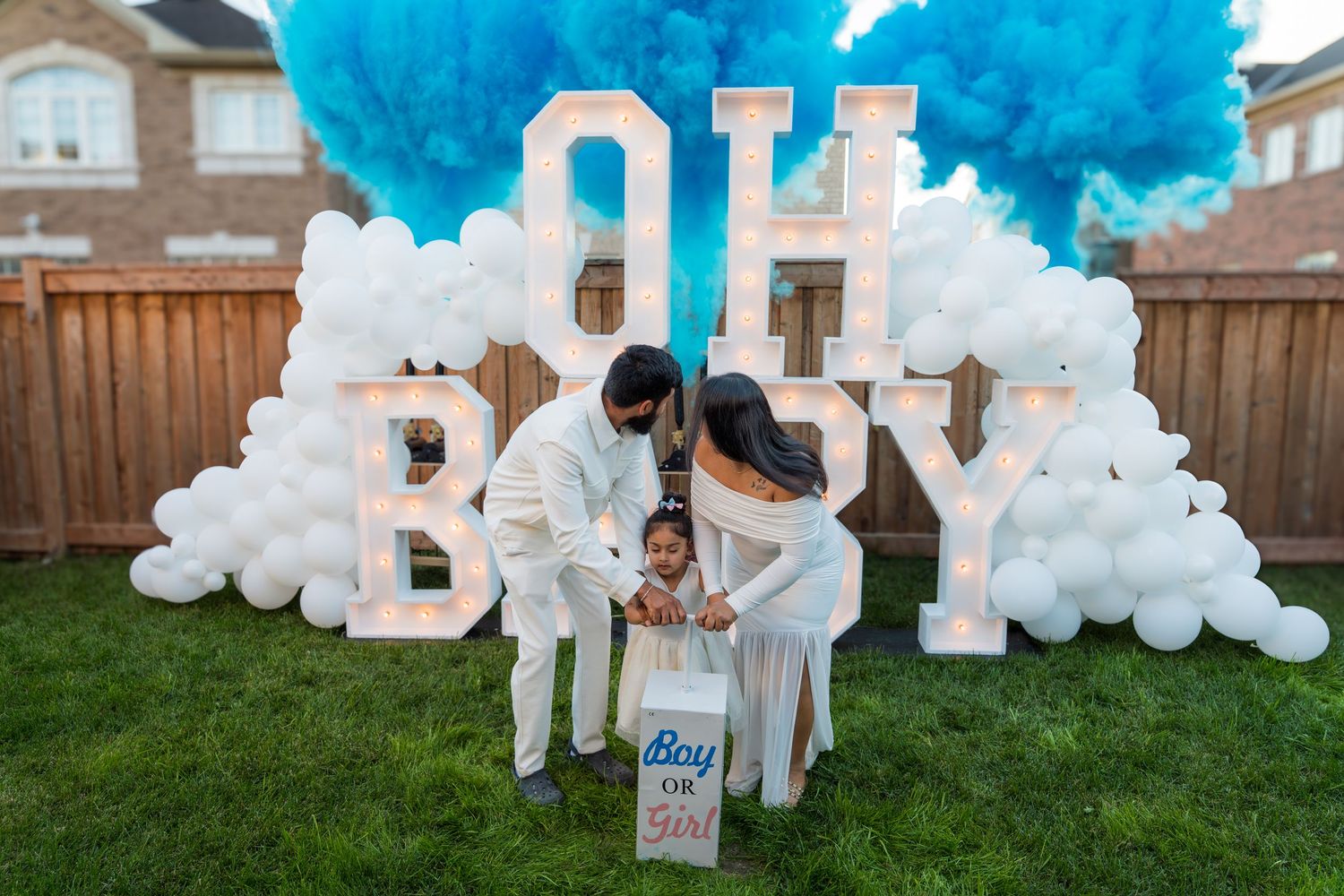 A couple reveals baby gender with blue smoke and OH BABY balloon backdrop in a backyard celebration.