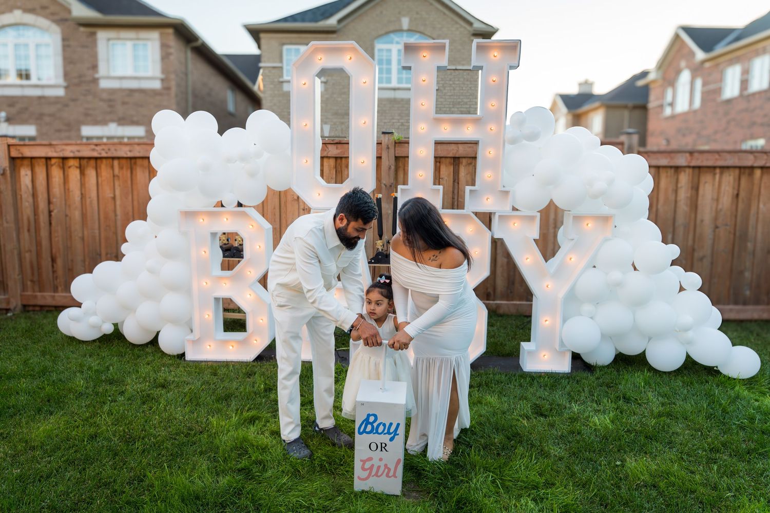 Family stands before an illuminated OH BABY sign surrounded by white balloon arch at a backyard gender reveal event.