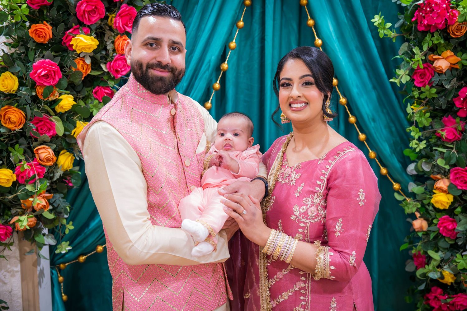 A couple in traditional South Asian attire holding their newborn baby in front of a colorful floral backdrop.