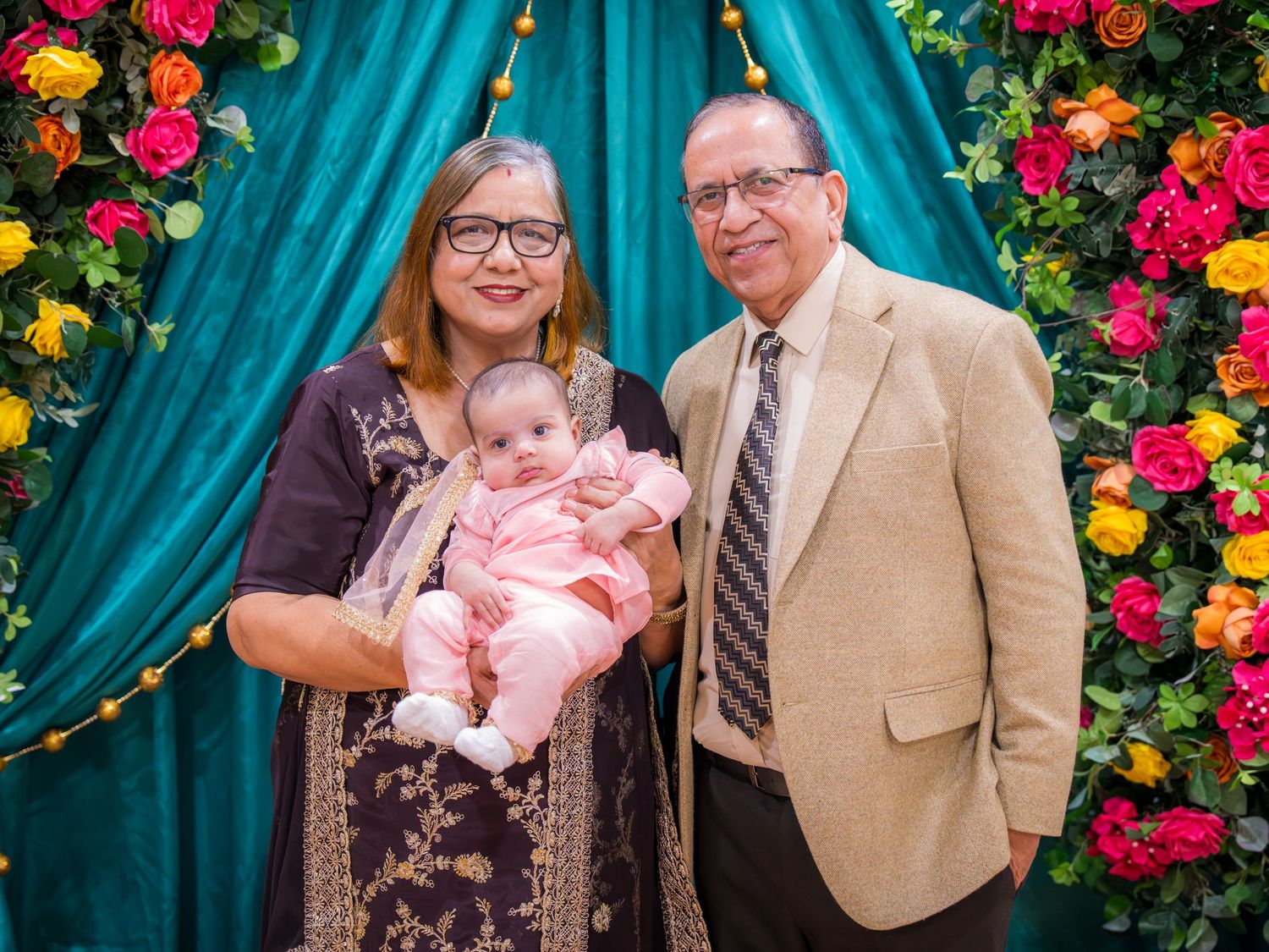 Grandparents posing with a baby in front of colorful floral backdrop and teal curtains at a family celebration.