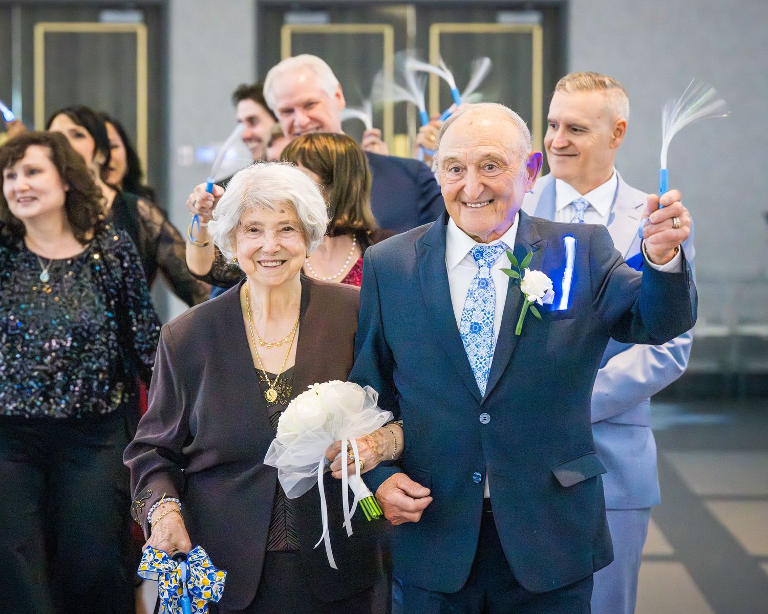 Elderly couple walks through cheering guests holding light wands at anniversary celebration.