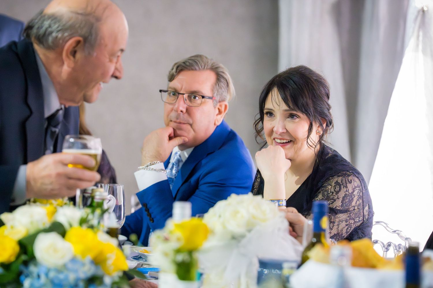 Three elegantly dressed guests conversing and smiling at a formal banquet table with yellow floral arrangements.
