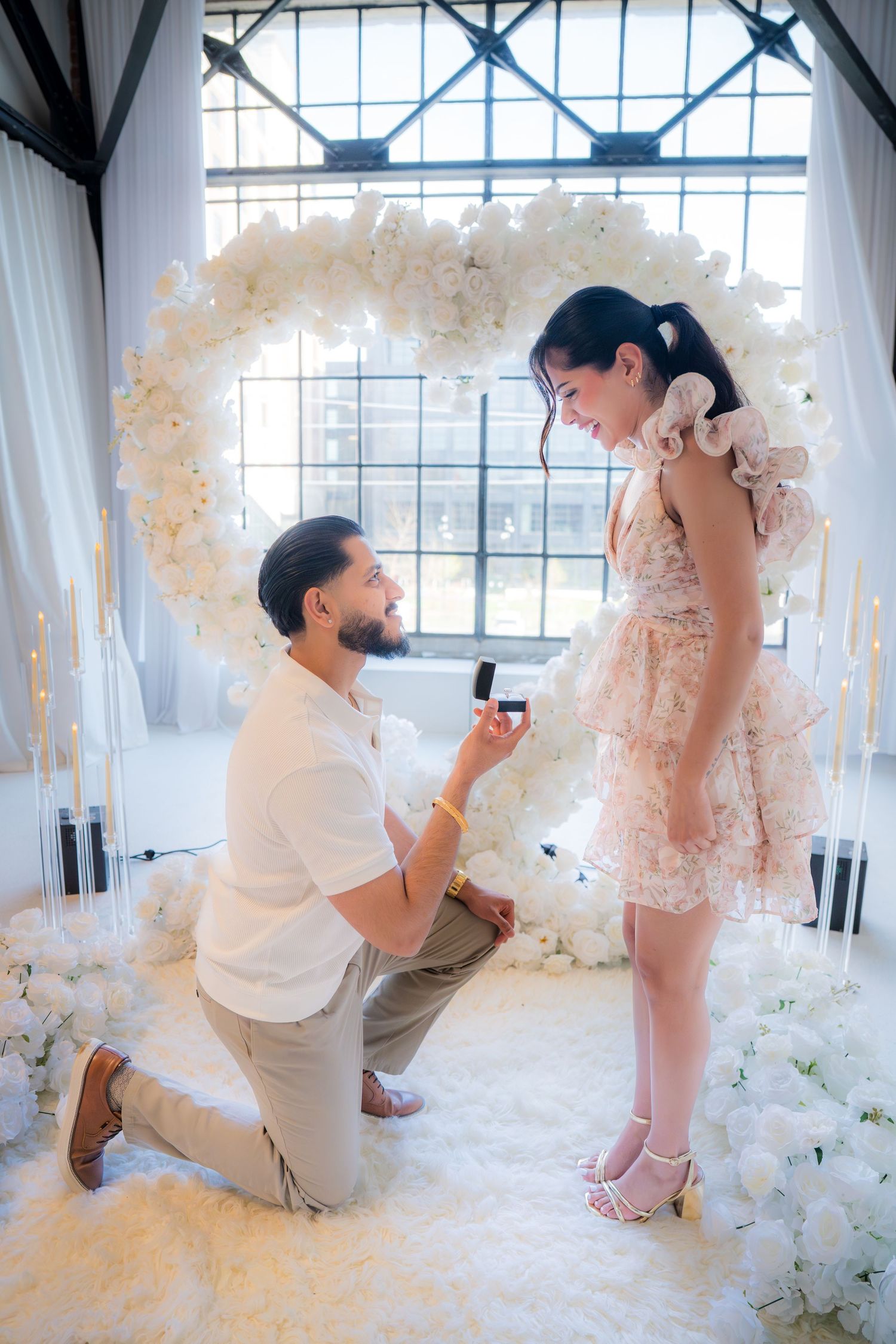 Man on one knee proposing to woman in floral dress in front of white heart floral arch.