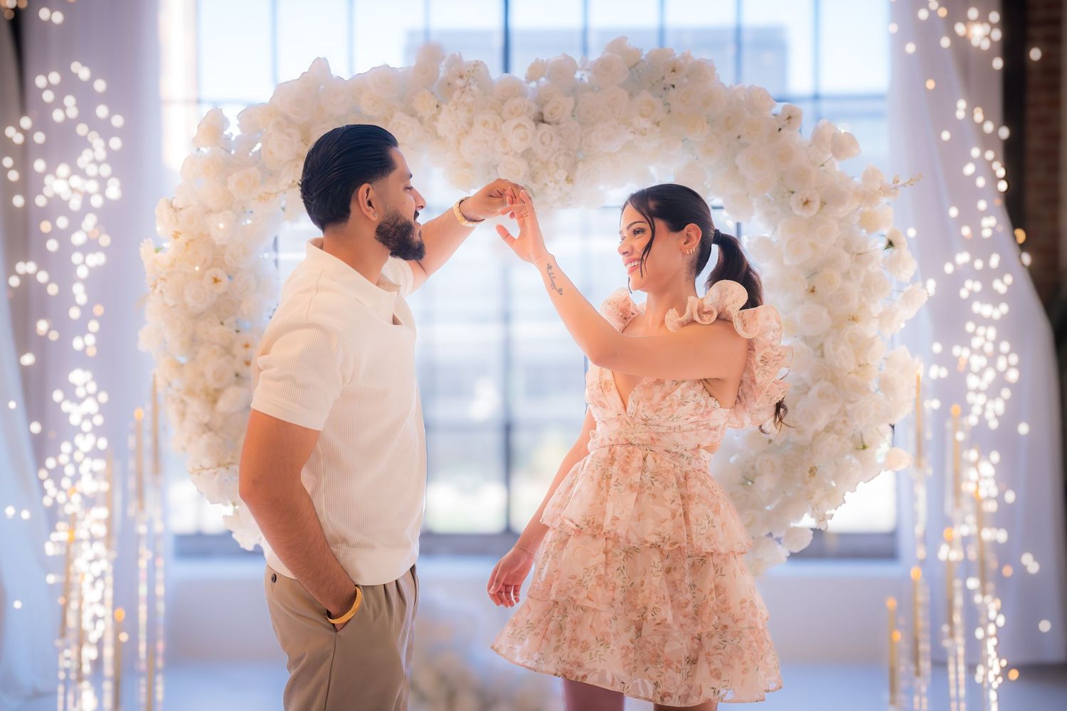 Couple dancing joyfully in front of a illuminated white floral heart arch during a romantic proposal.