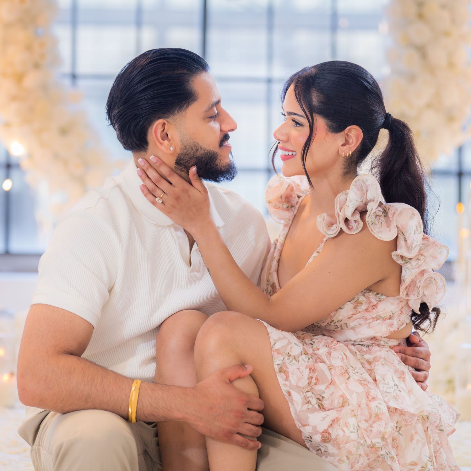 Couple sharing a romantic moment, woman in floral dress touching man's face, surrounded by white floral decor.